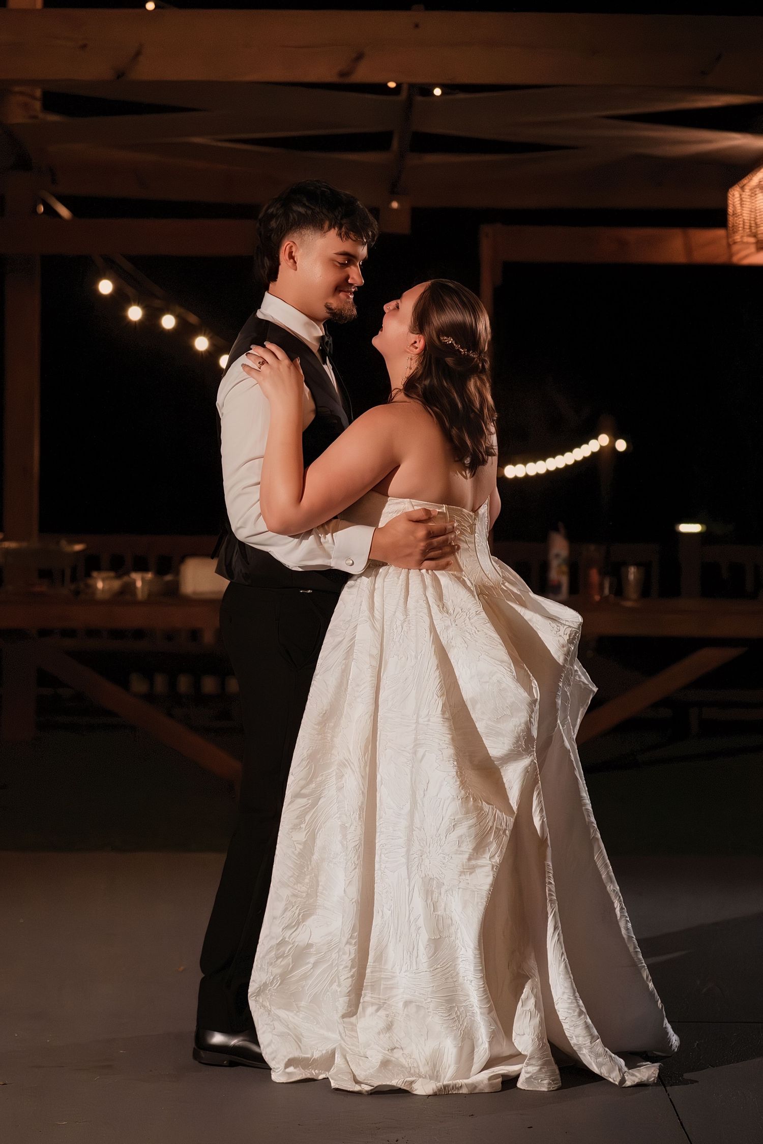 Bride and groom slow dance together under warm string lights at elegant outdoor evening wedding reception.