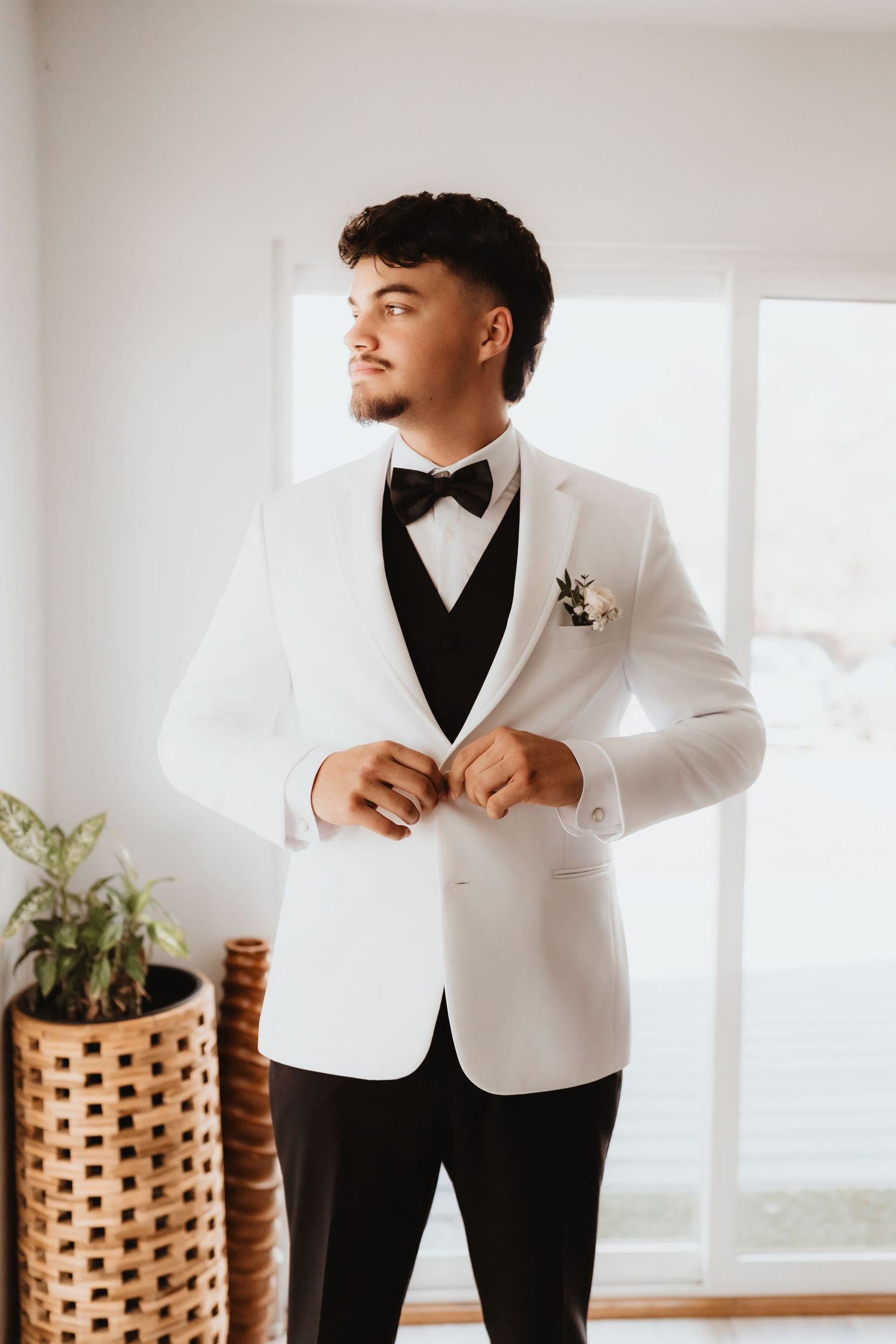 Groom in white tuxedo jacket with black bow tie and boutonniere buttons jacket near bright window.