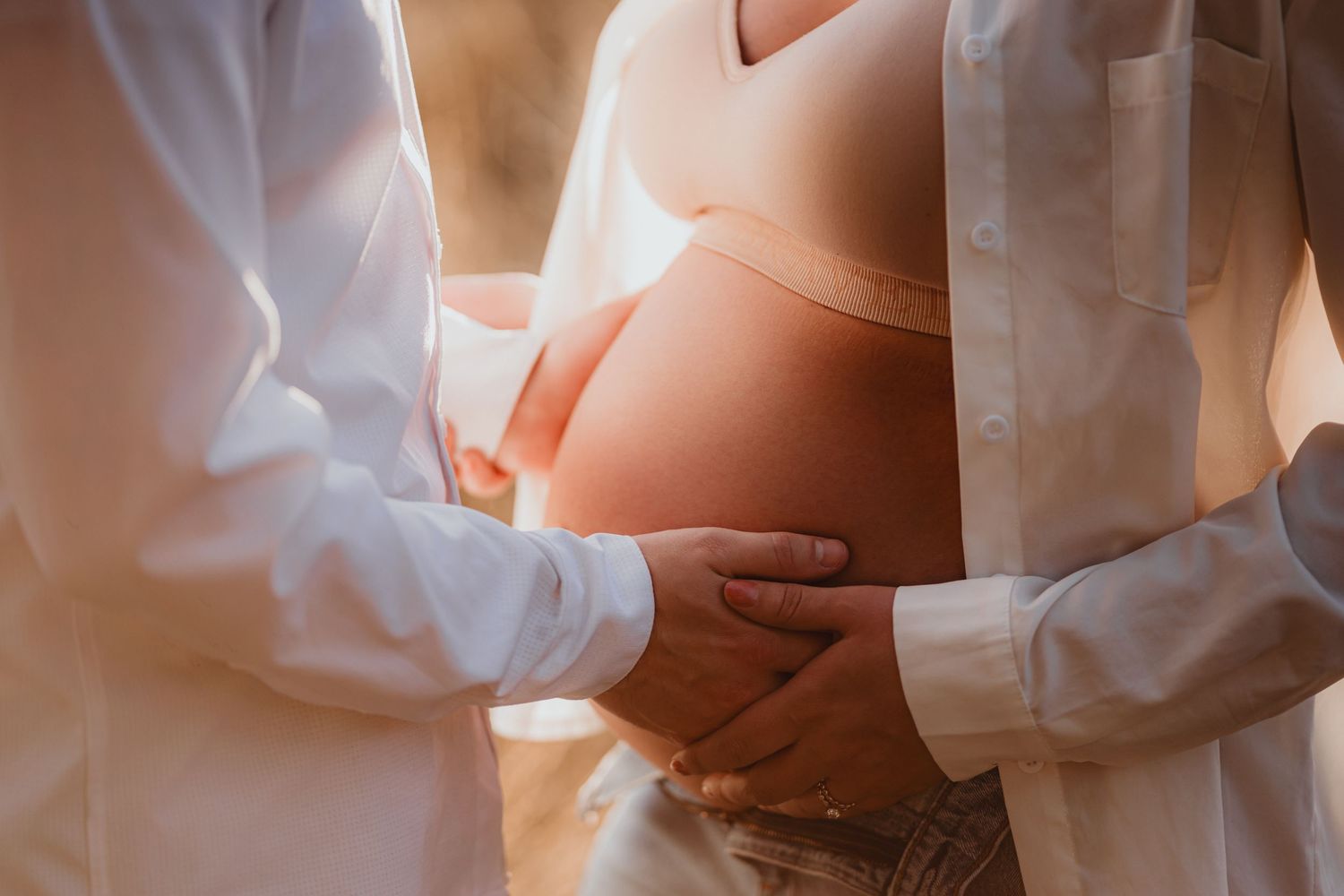 Couple tenderly cradling pregnant belly outdoors in warm golden light.