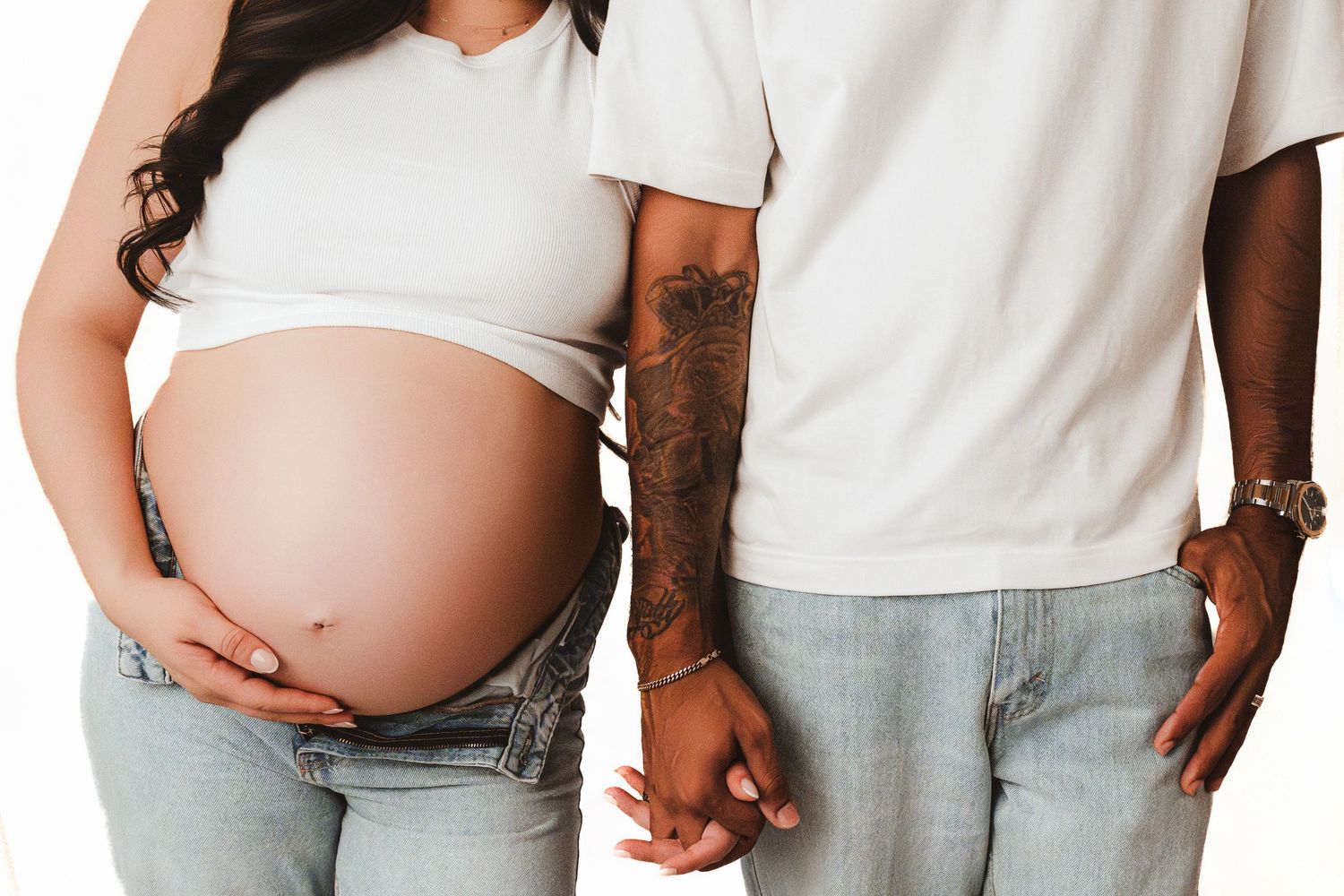 Close-up of pregnant couple holding hands, man with tattooed arm in white outfits and jeans.