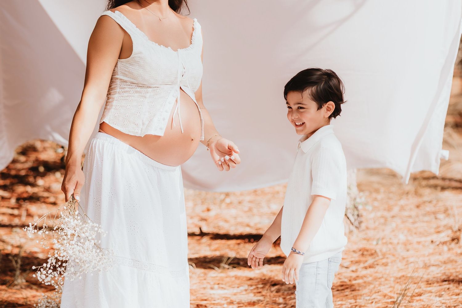 Smiling boy in white holding pregnant mothers hand outdoors during maternity photoshoot.