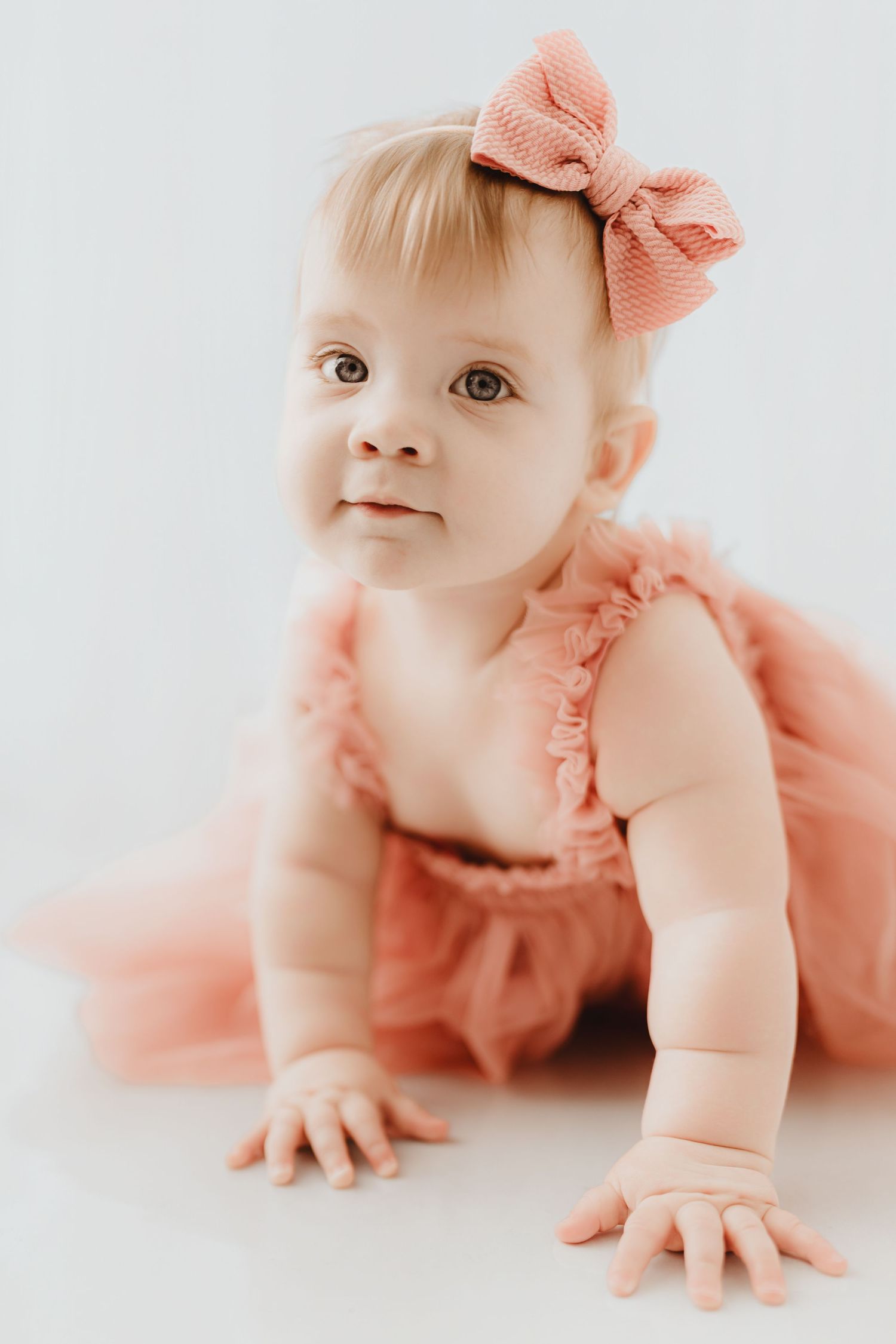 Sweet baby girl in pink ruffled romper and bow headband smiling while crawling on white background.