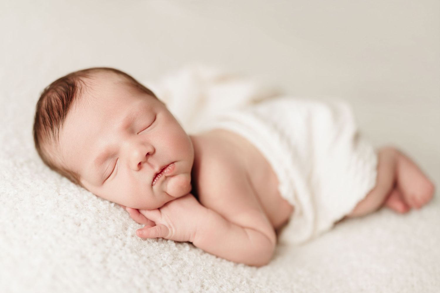 Newborn baby sleeping peacefully wrapped in white swaddle on soft cream blanket.
