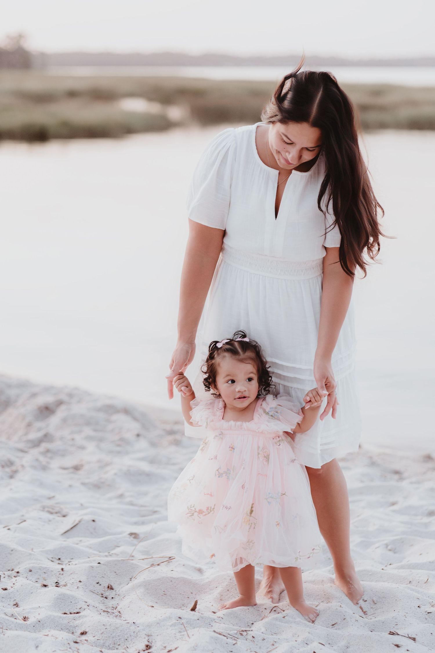 Mother in white dress holding toddler daughters hands on sandy beach at sunset.