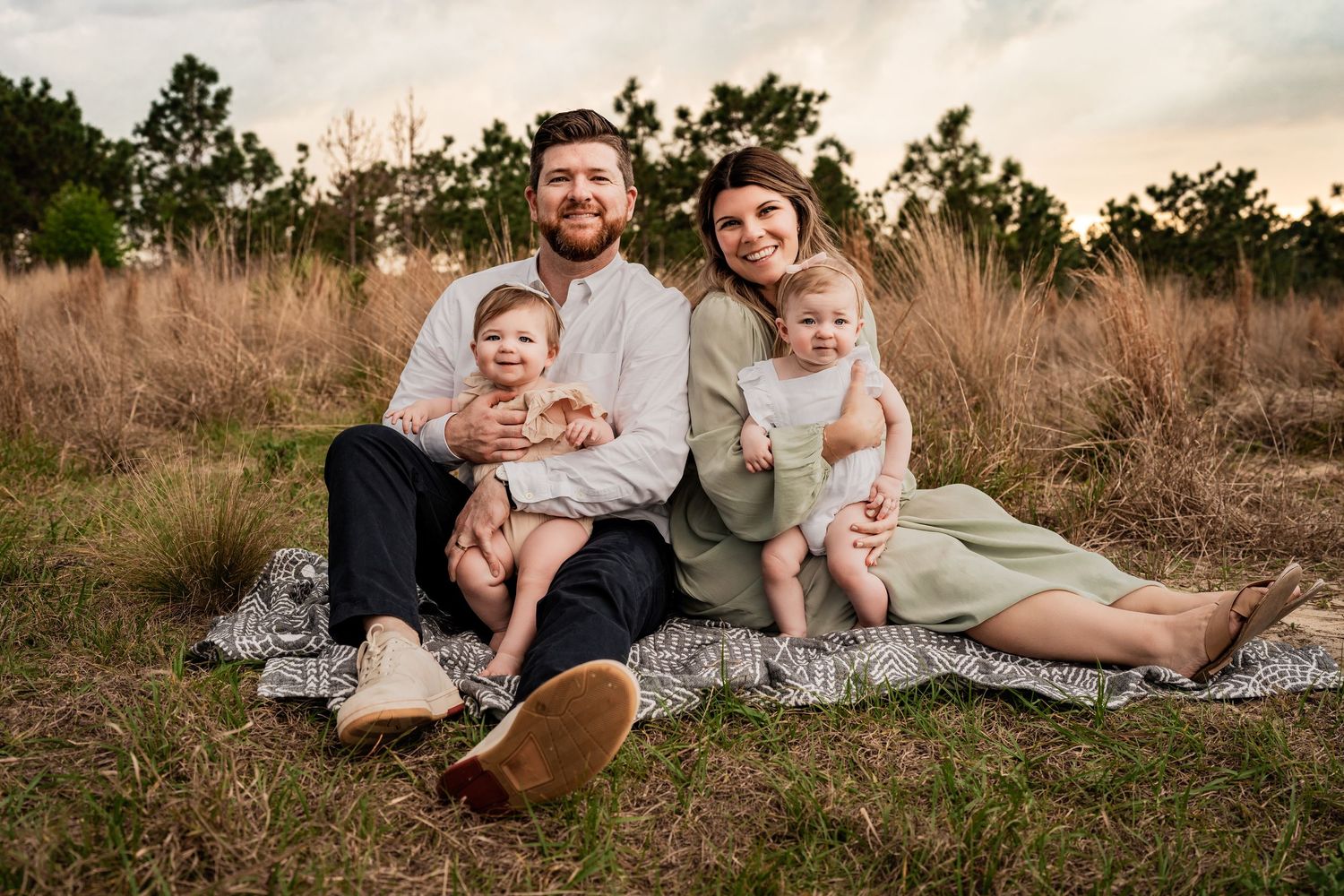 Happy family of four with twin babies sitting on a blanket in a golden field during sunset portrait.