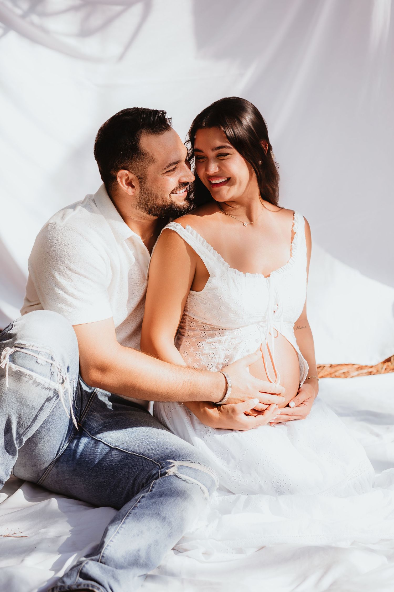 Smiling couple in white outfits cradling pregnant belly during intimate maternity photo session.