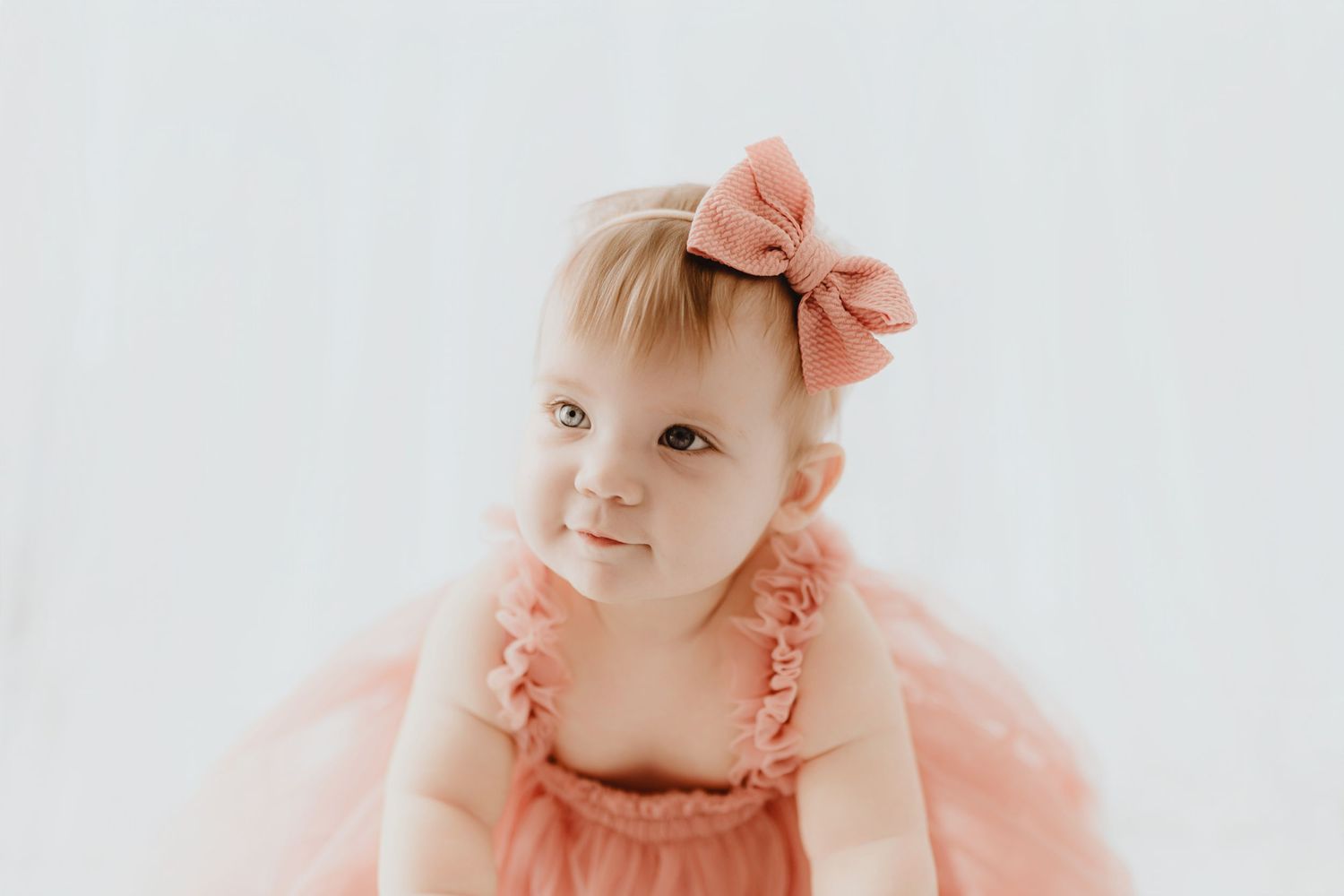 Sweet baby girl in pink ruffle dress and matching bow headband smiling softly.