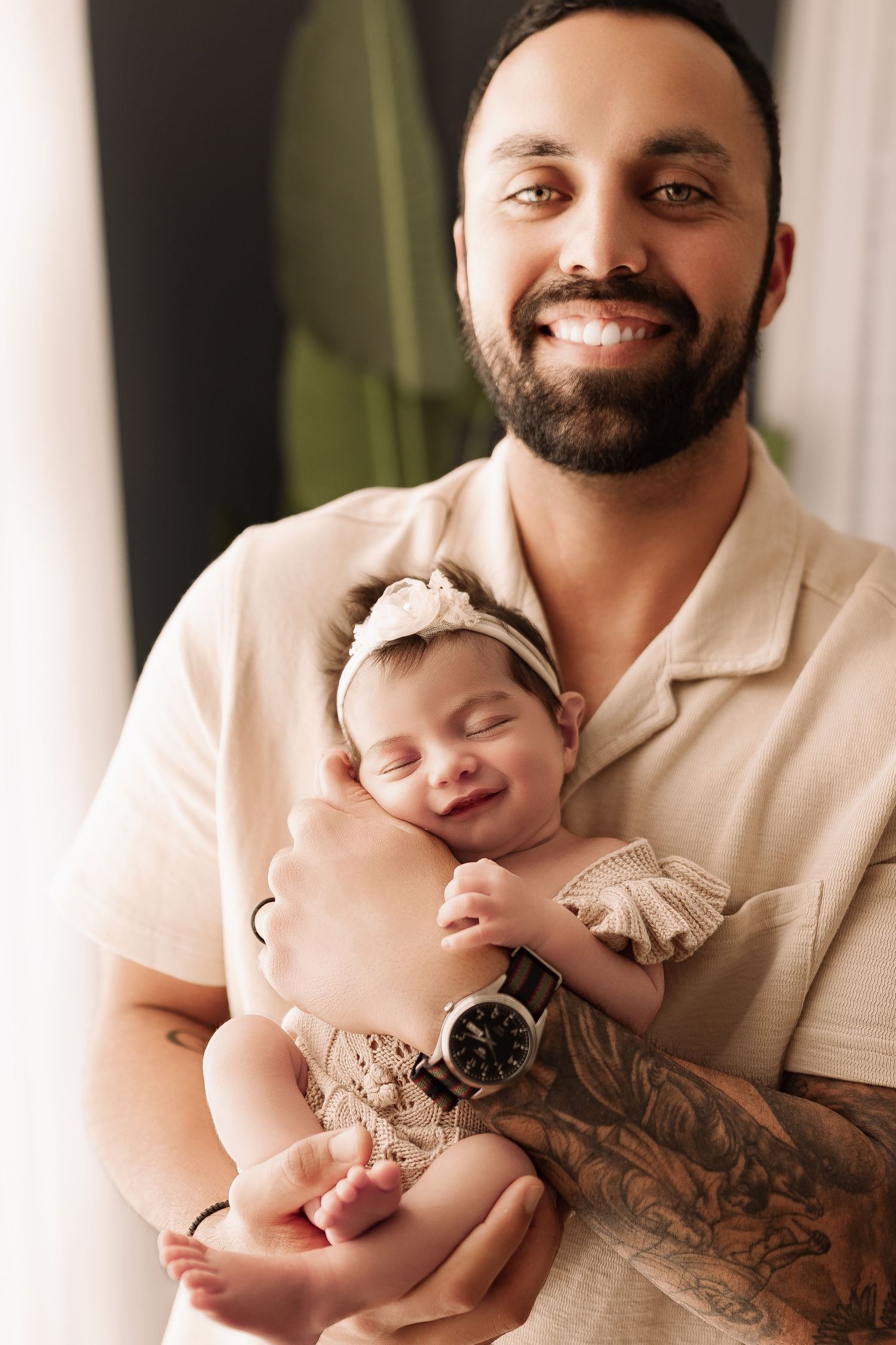 Smiling bearded father with tattoos holding sleeping newborn baby girl wearing a floral headband.