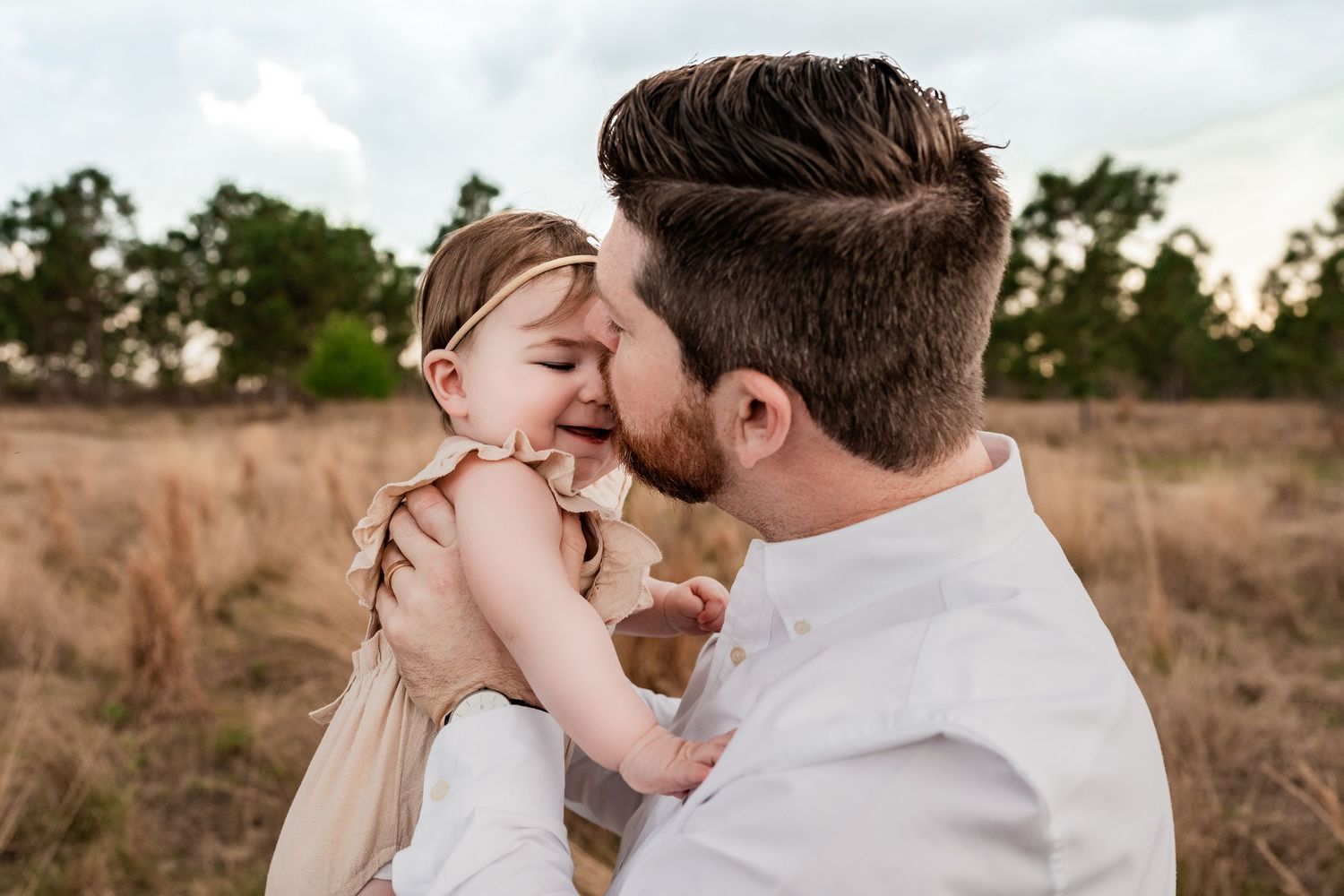 Father in white shirt kissing laughing baby girl in tan dress in a rustic outdoor field.