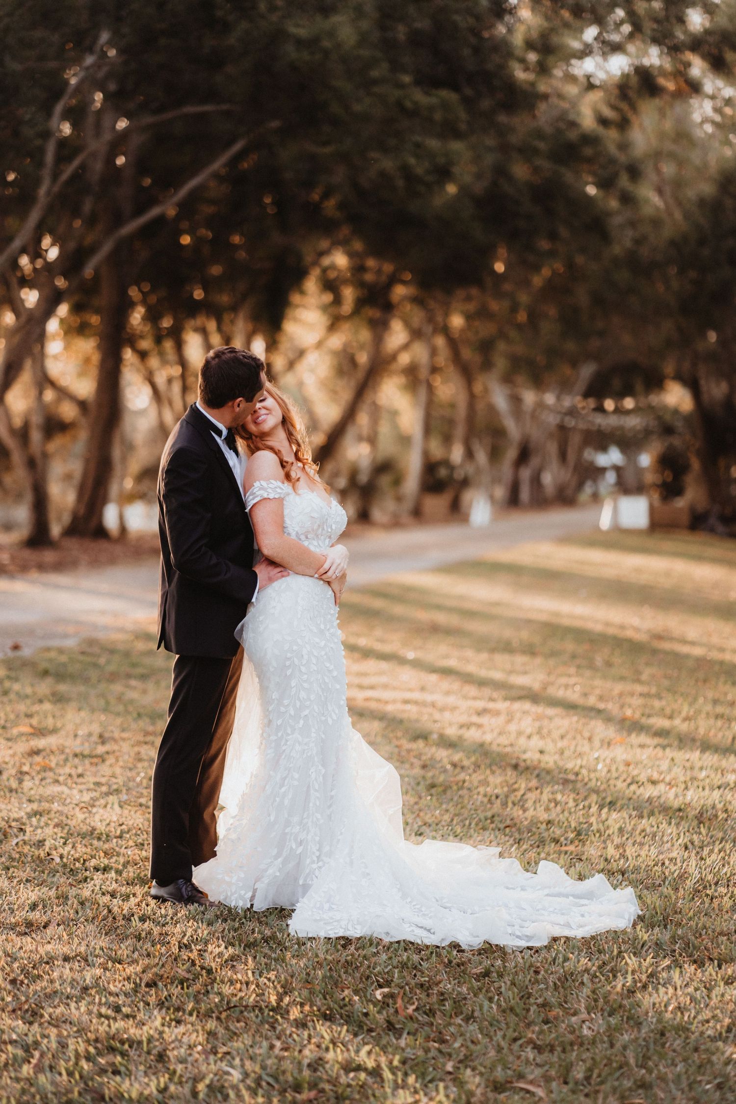 Newlyweds embrace in golden hour light among trees, bride in off-shoulder lace mermaid gown with long train.