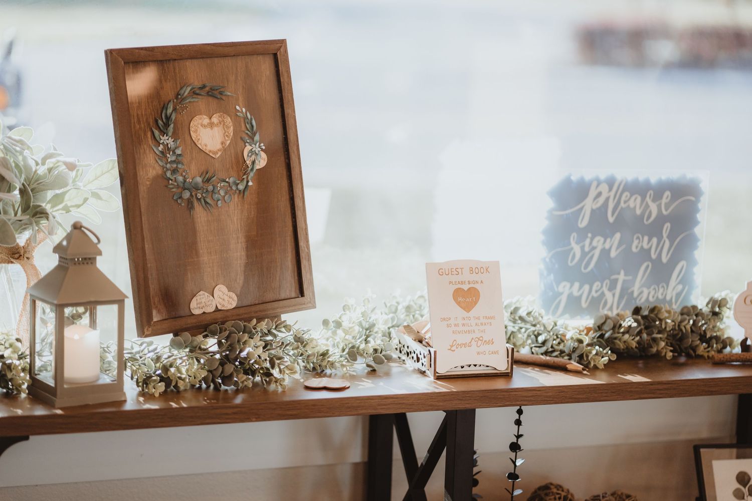 Rustic wedding guest book table with wooden heart drop frame, lantern, and greenery decor.