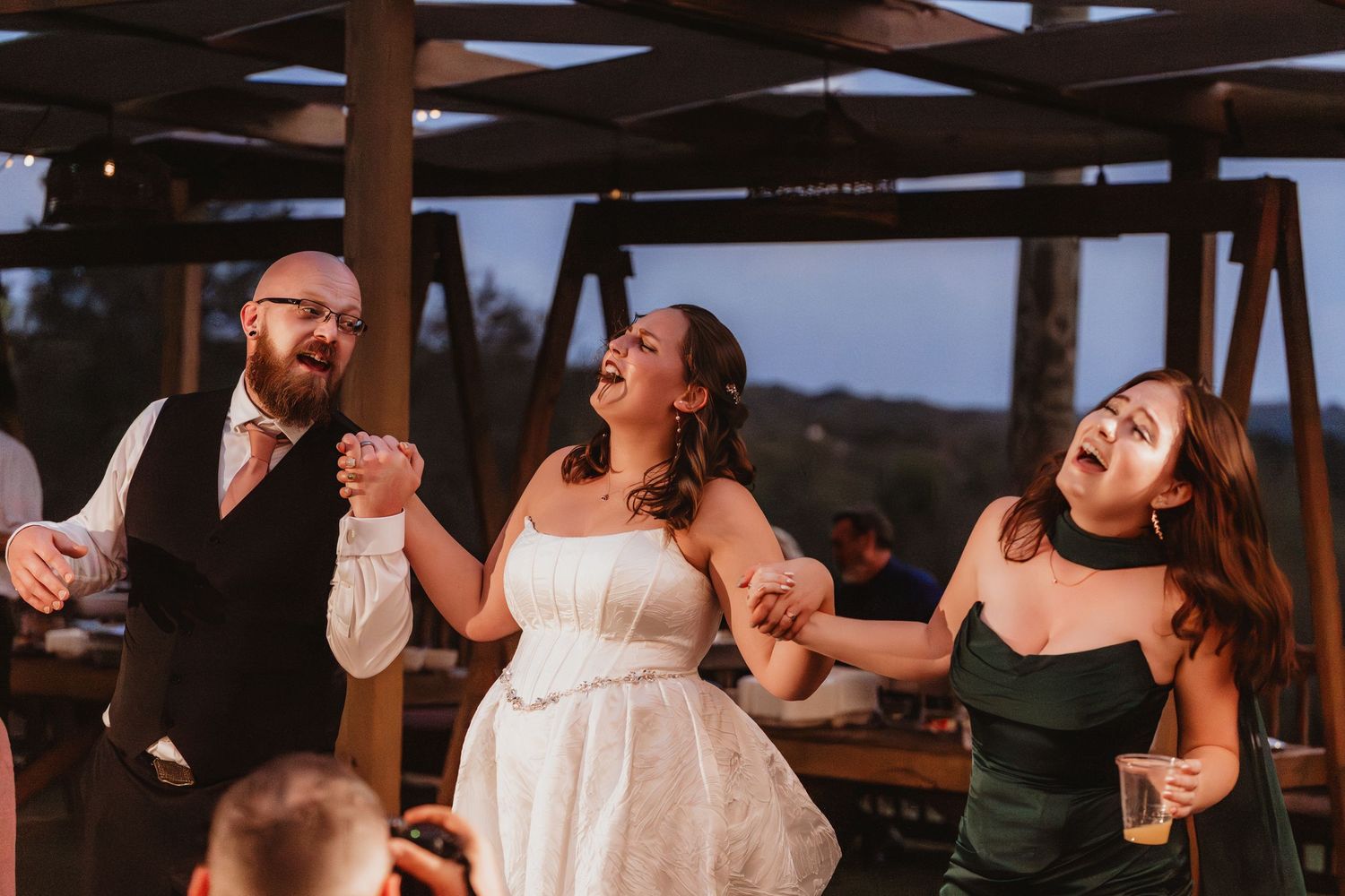 Joyful wedding reception dancing with bride in white gown laughing alongside groom and bridesmaid in green dress.