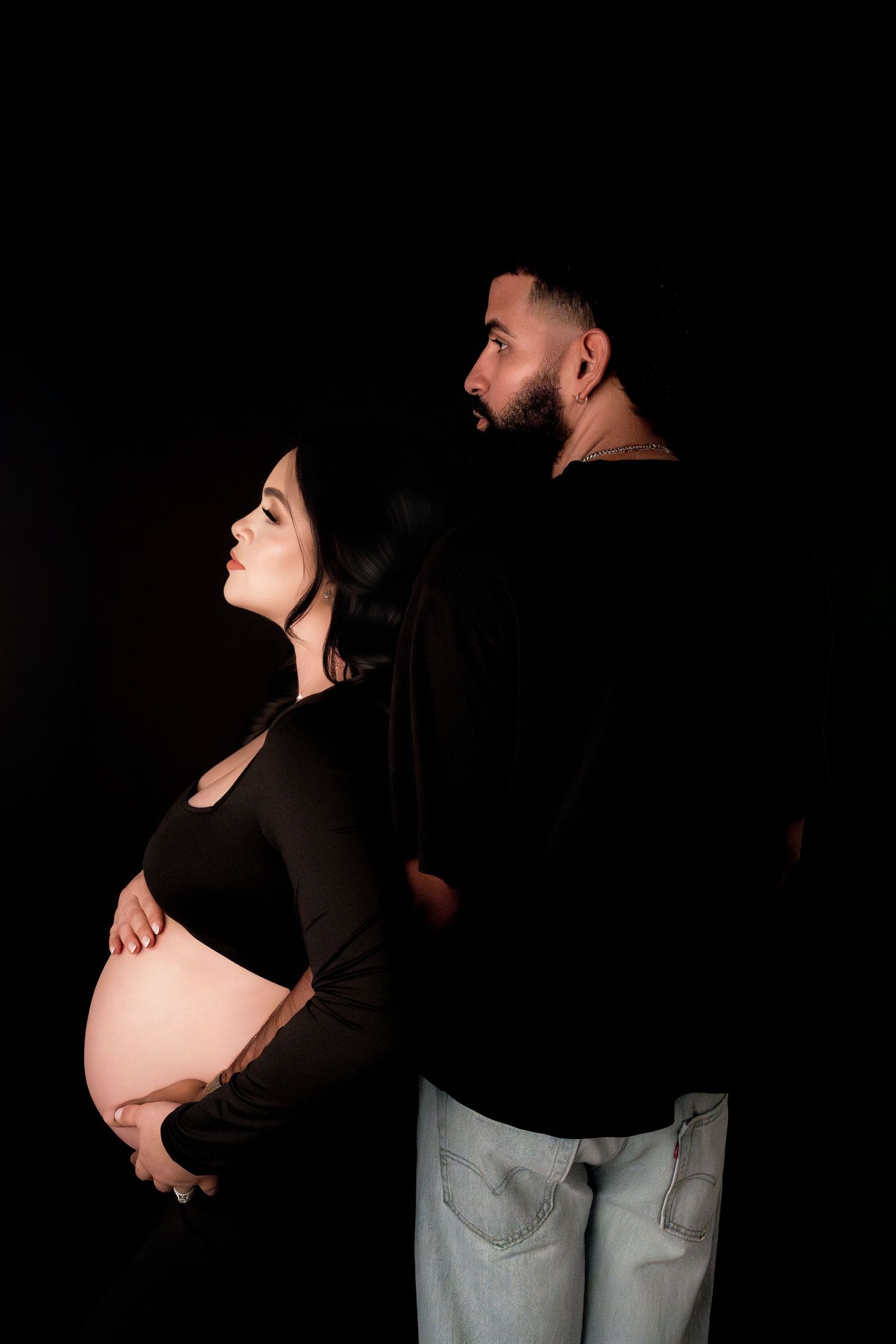 Pregnant couple posing back-to-back in black outfits against dark background, woman cradling baby bump.