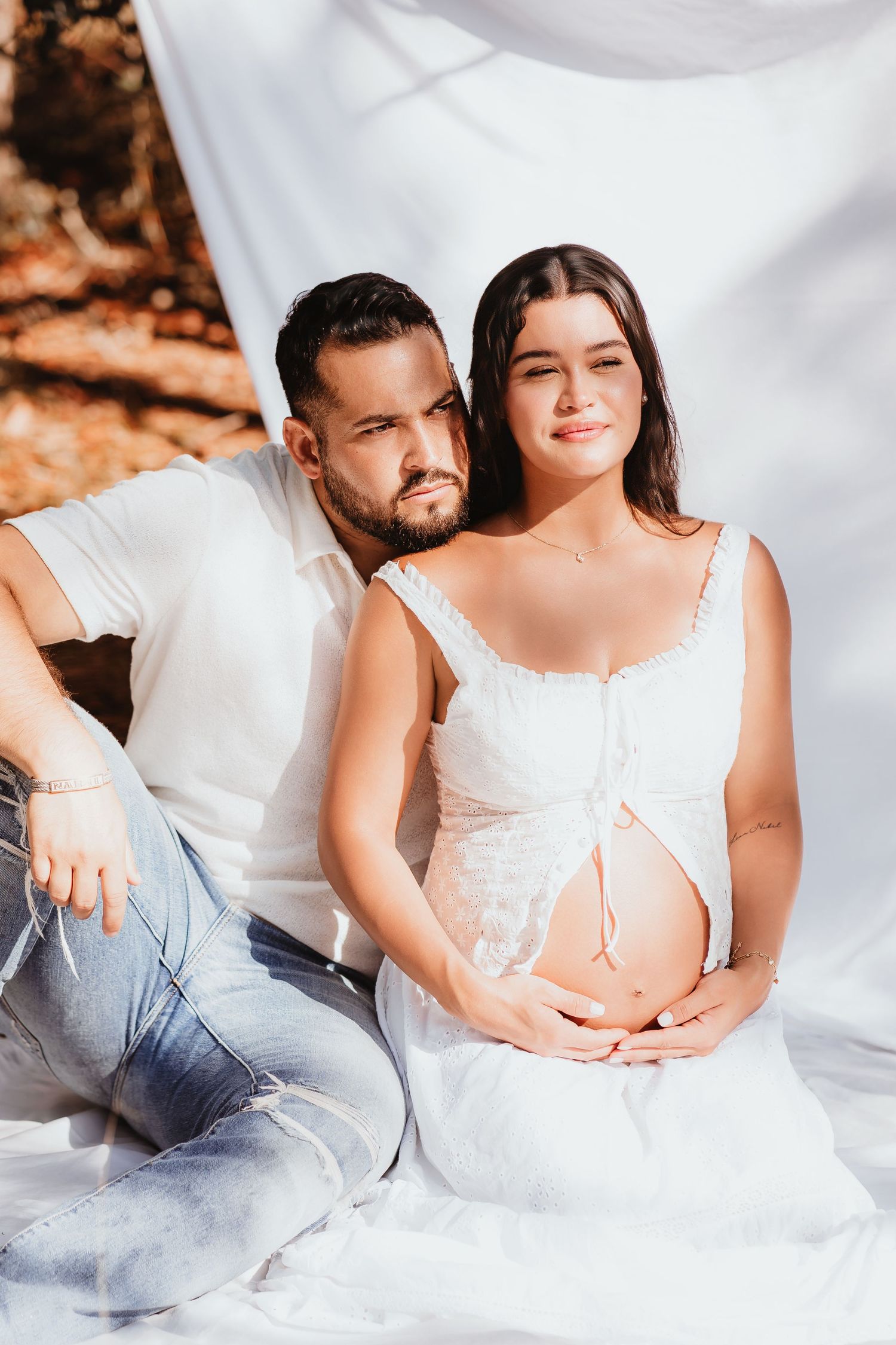 Couple posing together with pregnant woman in white dress cradling her baby bump.