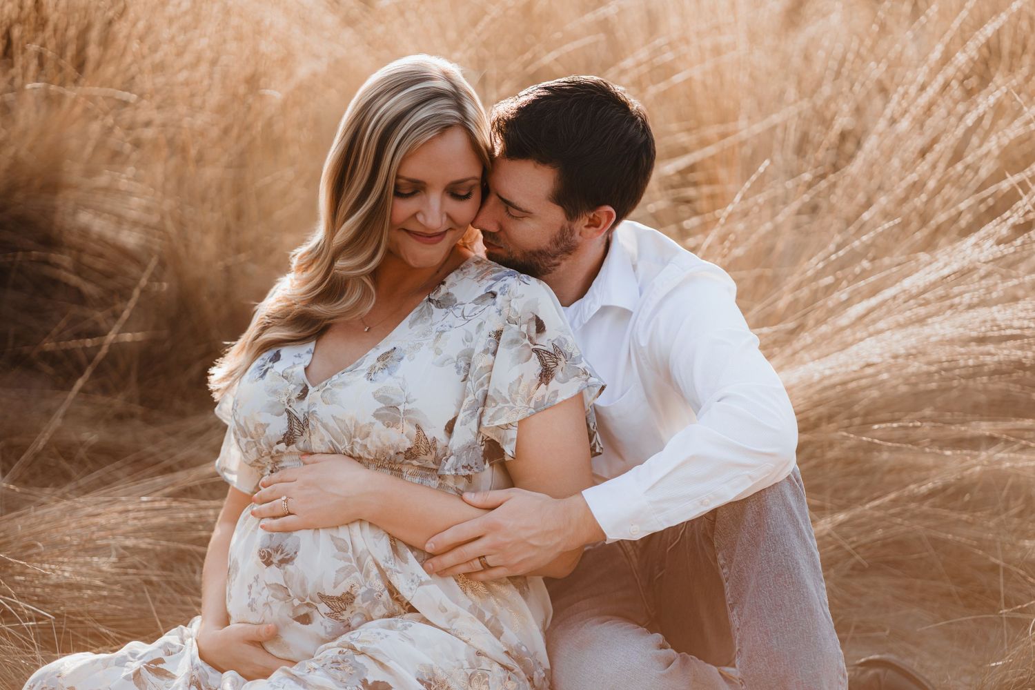 Couple sits intimately in tall golden grass, man nuzzling pregnant partner during outdoor maternity session.