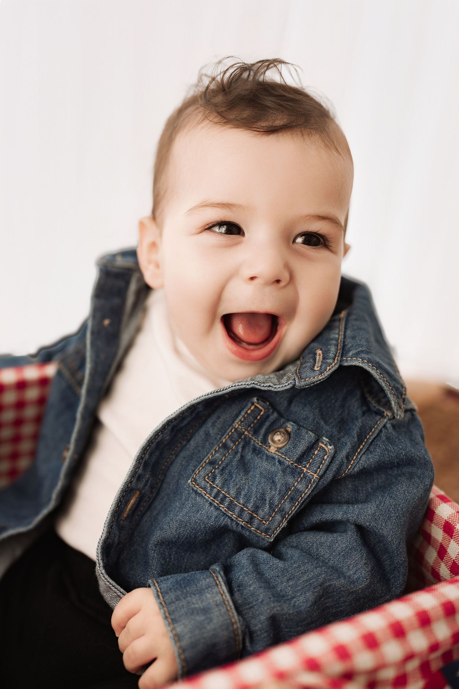 Happy laughing baby wearing a denim jacket sitting in a red checkered picnic basket.