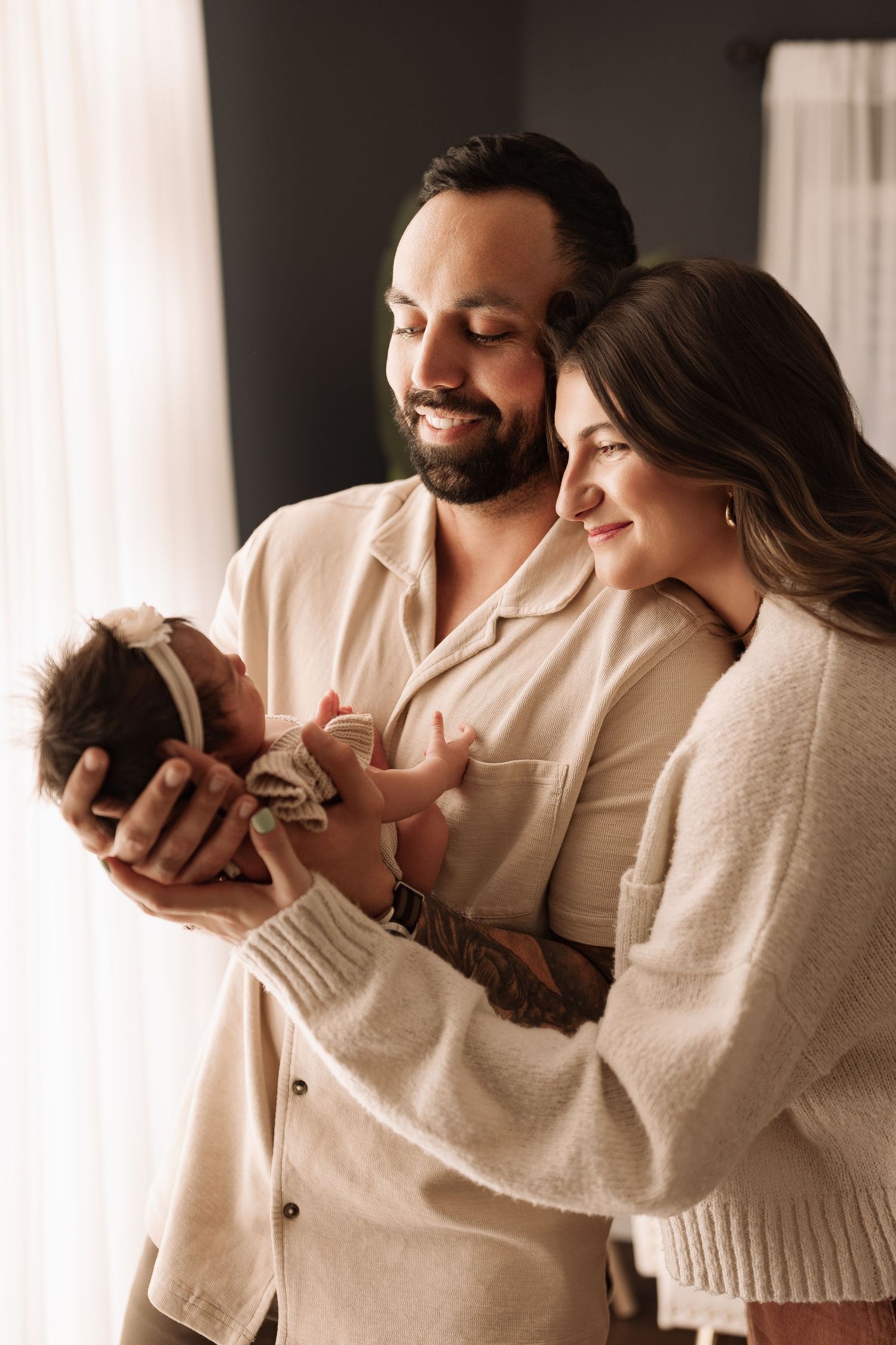 Smiling parents hold their newborn baby girl together near a sunlit window at home.