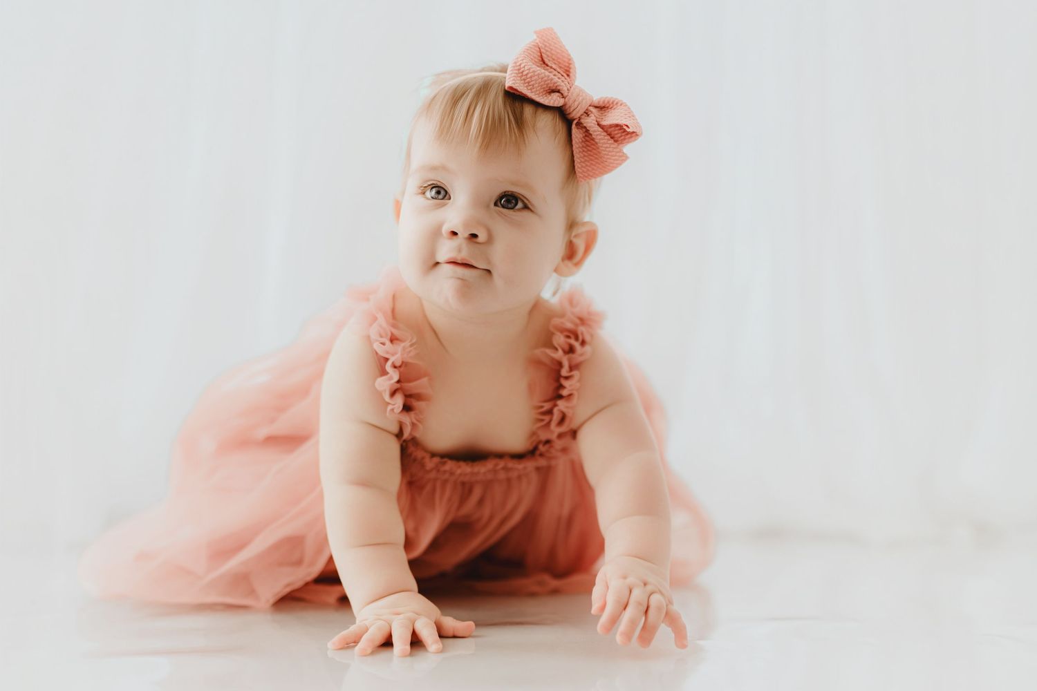 Sweet baby girl in dusty rose tulle dress and bow headband crawling forward.