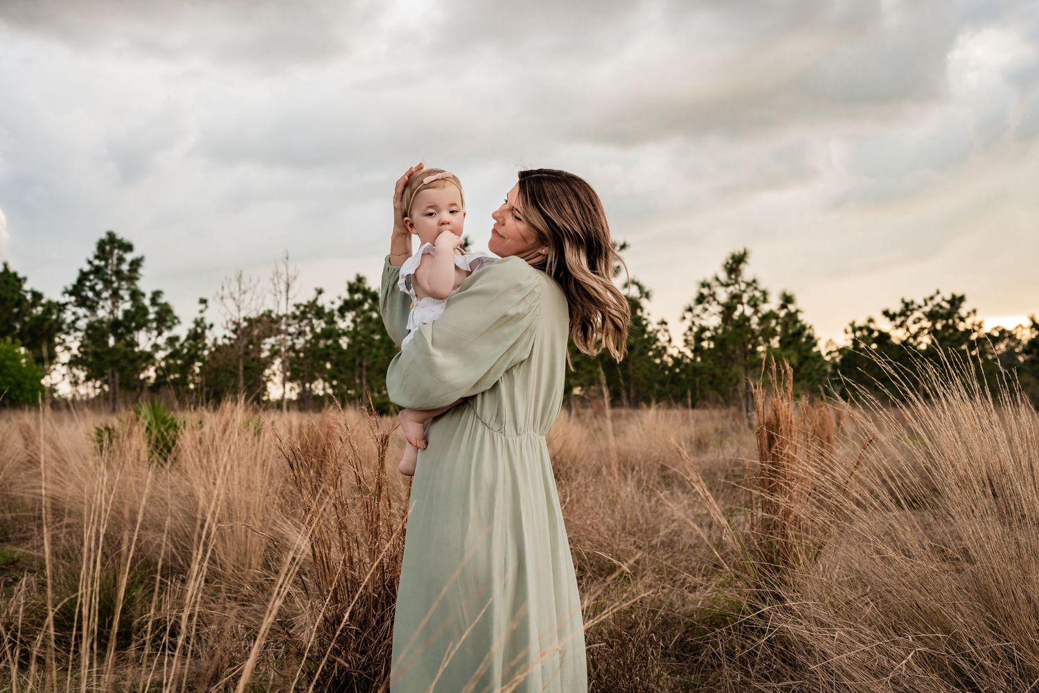 Mother in sage green dress holding baby girl while standing in tall golden grass at sunset.