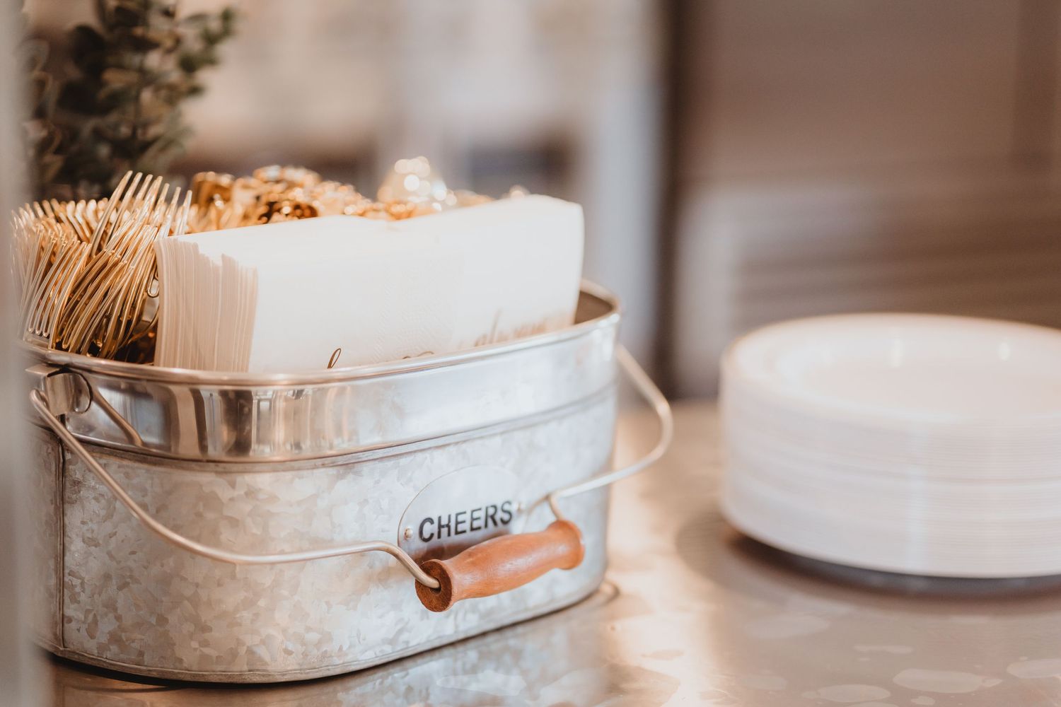 Galvanized metal bucket labeled 'Cheers' holding napkins and toothpicks beside white plates at a wedding reception.