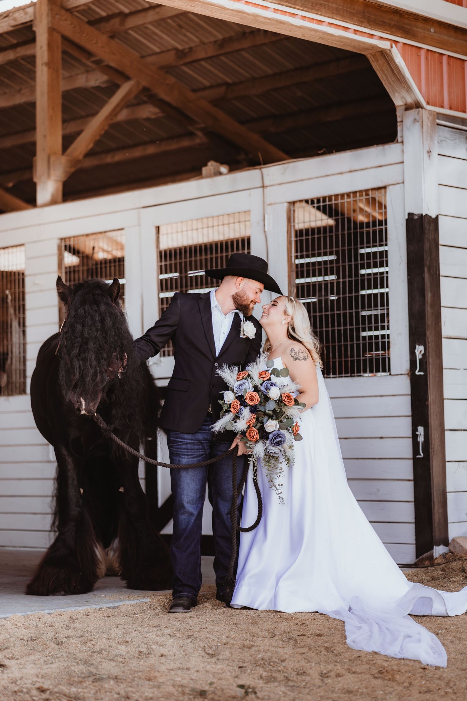 Western-themed bride and groom kiss beside a black horse at barn stables.