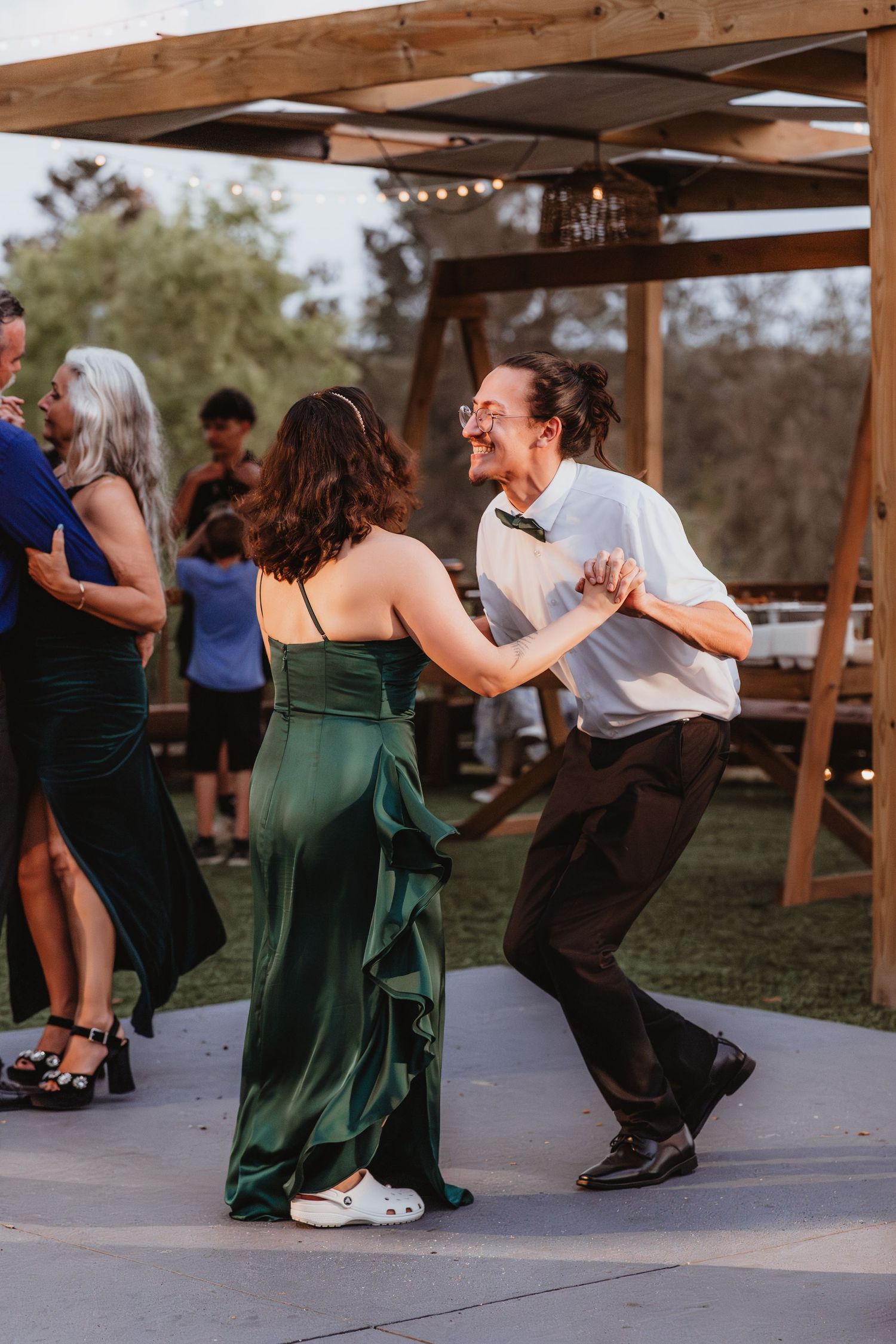 Couple in green and white dancing energetically at outdoor wedding reception under string lights.