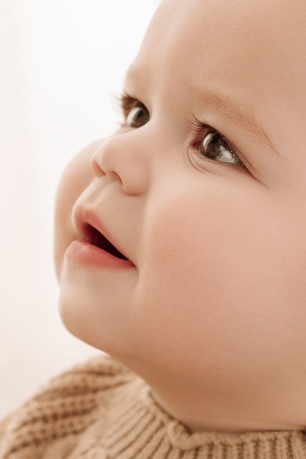 Close-up portrait of an adorable smiling baby with brown eyes wearing a knit sweater, looking up with curiosity.