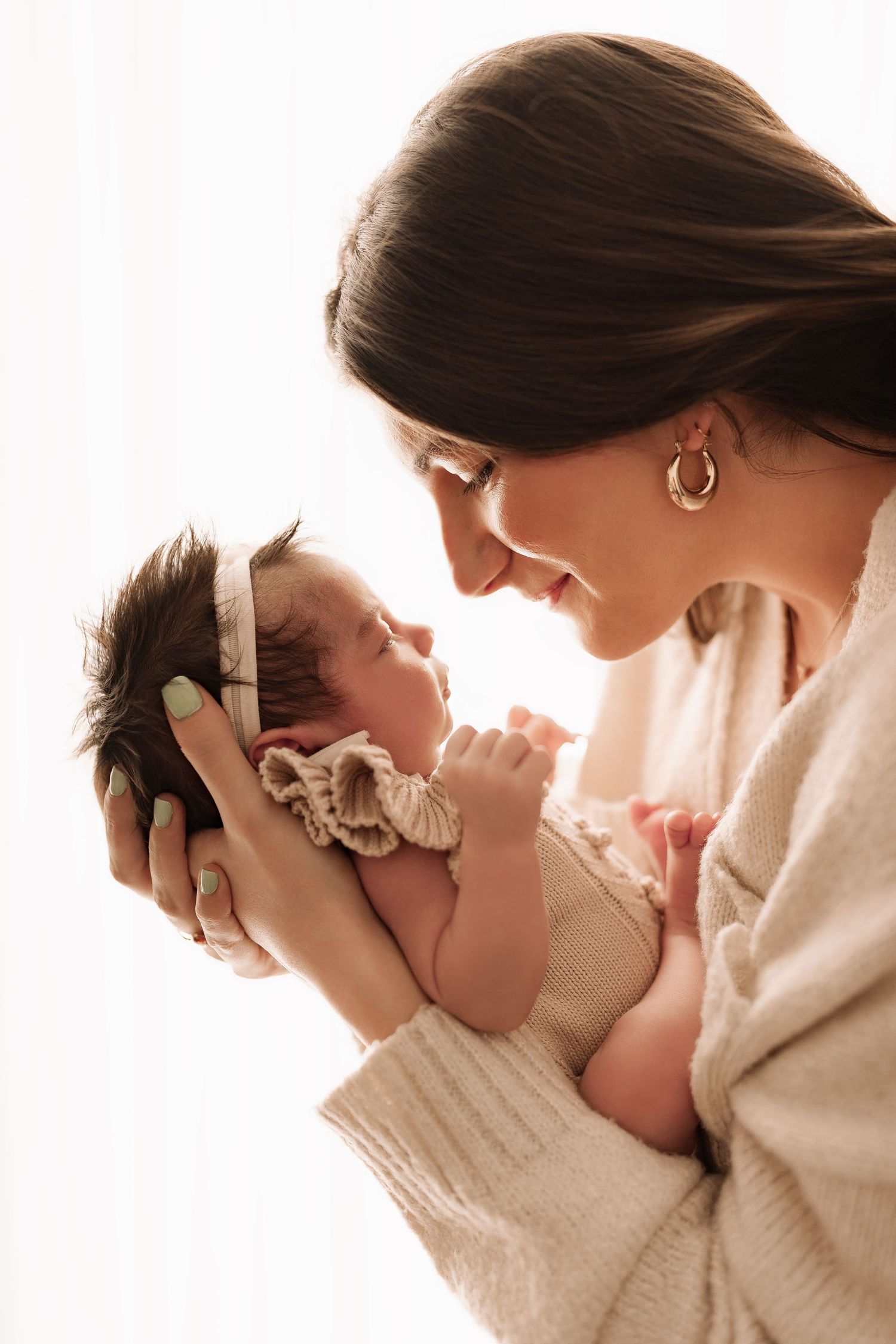 A smiling mother gazes lovingly at her newborn baby girl wearing a white headband.