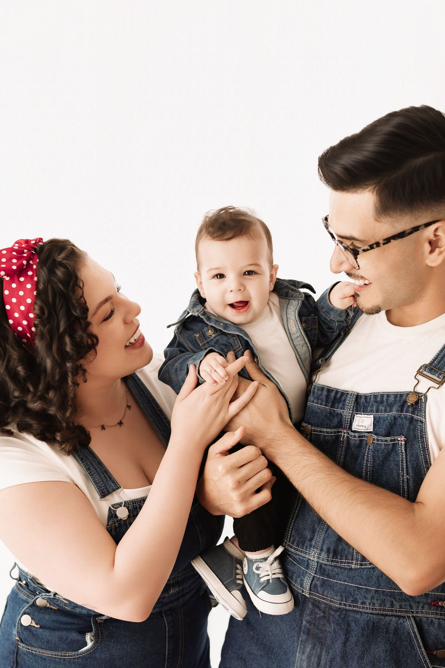 Loving parents in denim overalls smiling and playing with their happy baby on white background.