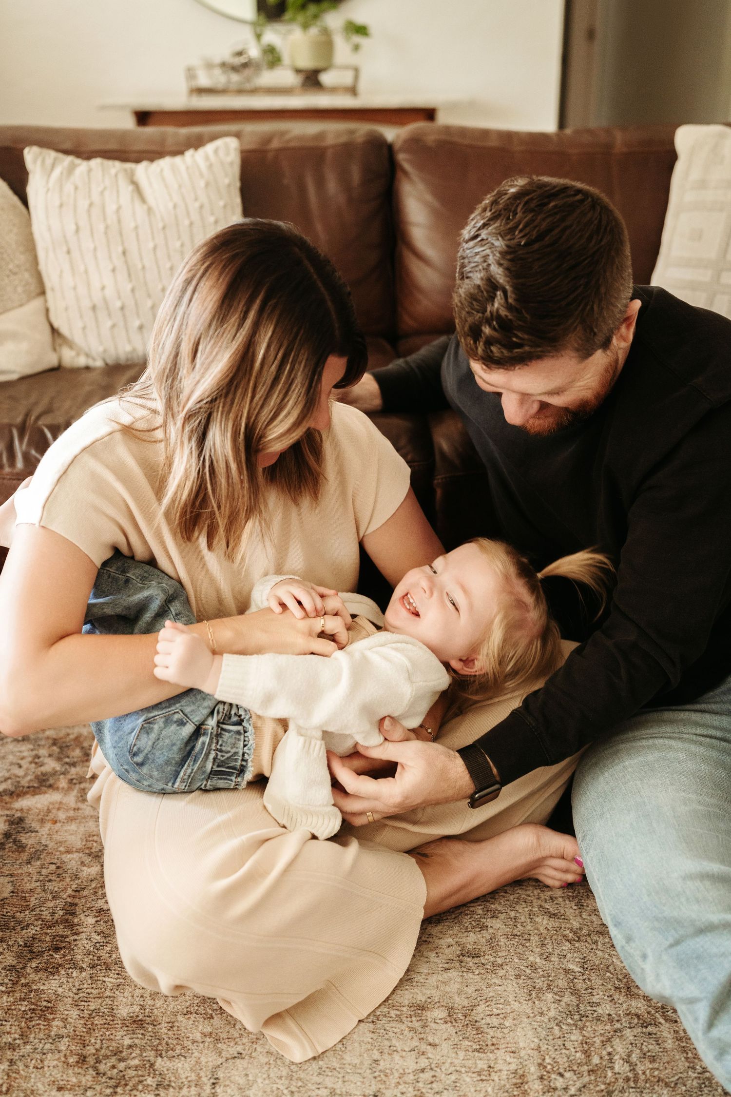 Loving parents playfully tickling their laughing toddler on a soft carpet in a warm living room.