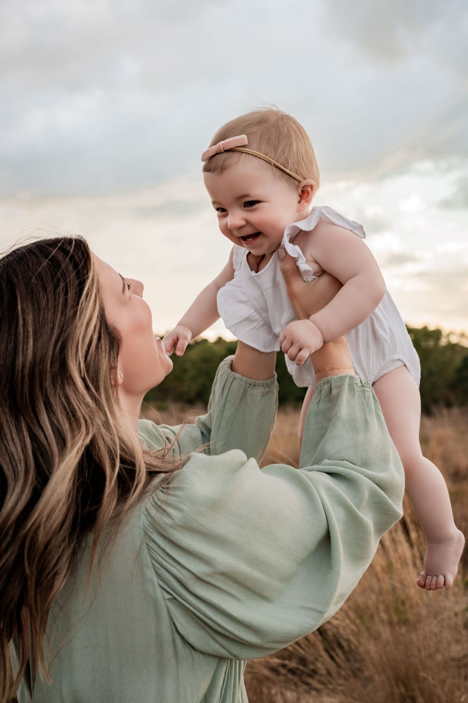 Mother in sage green dress lifts laughing baby girl in white outfit against golden field backdrop.