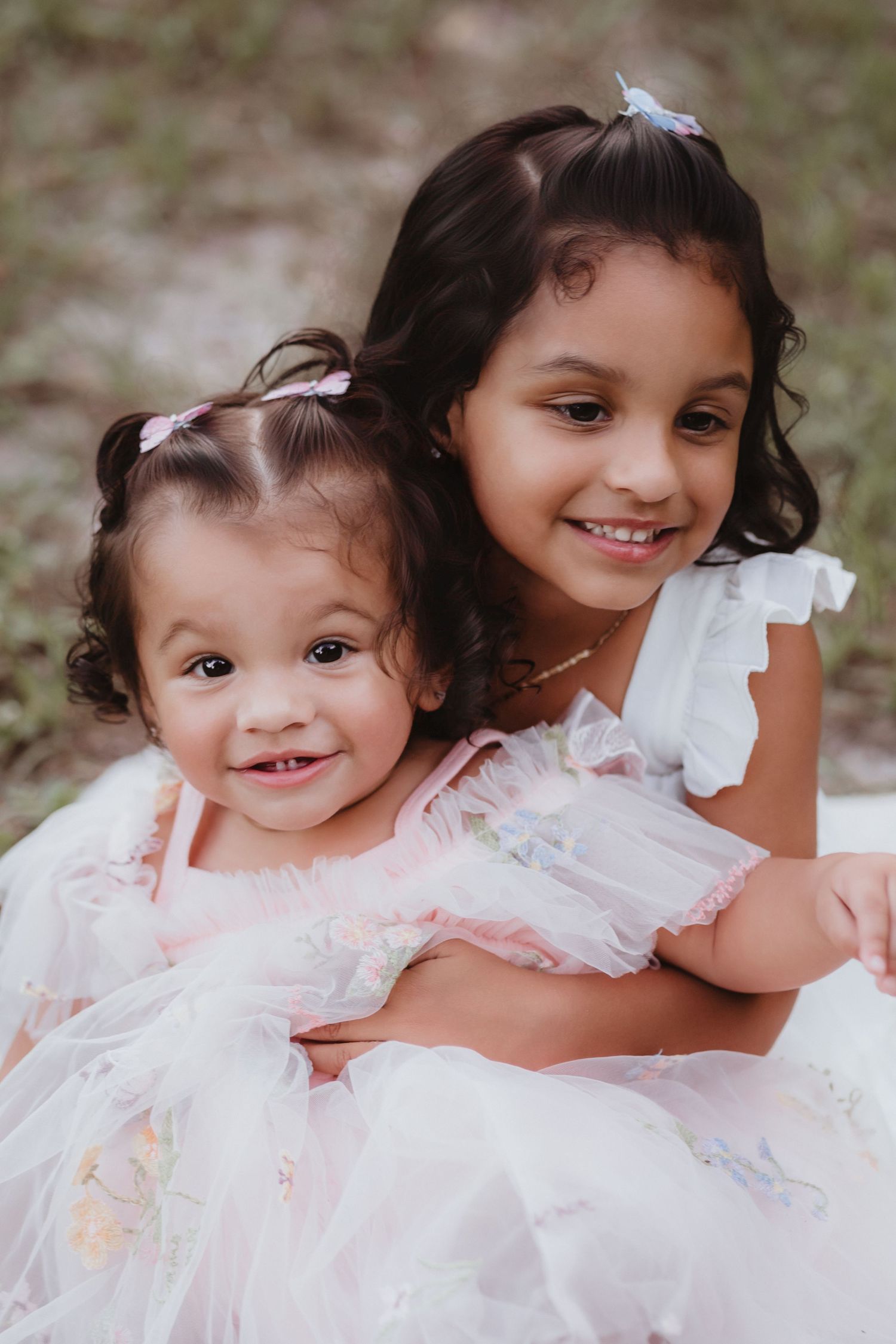 Two young sisters in white and pink dresses sitting together and smiling outdoors.