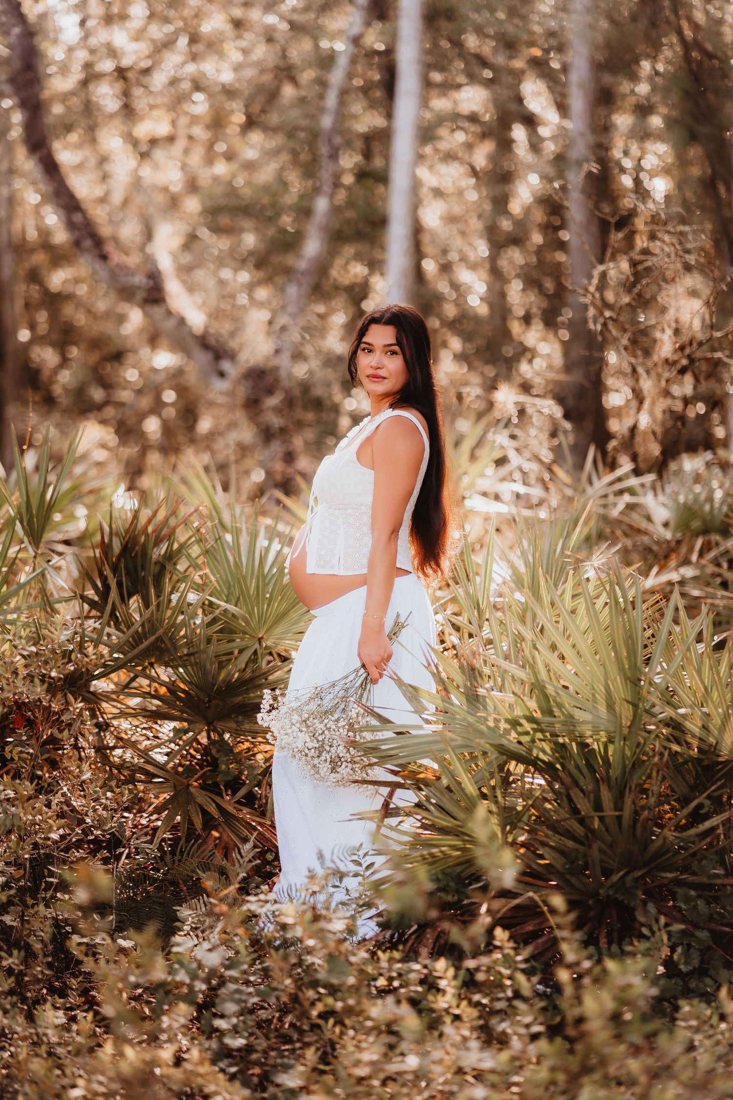 Pregnant woman in white outfit standing among palm plants in golden forest light.