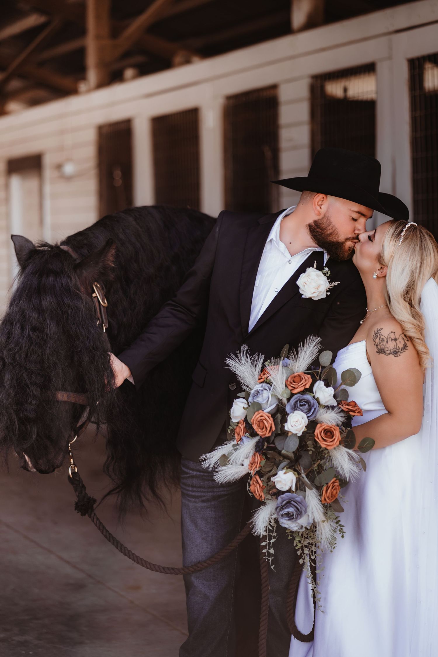 Newlyweds share a kiss beside a black horse in a stable barn setting.