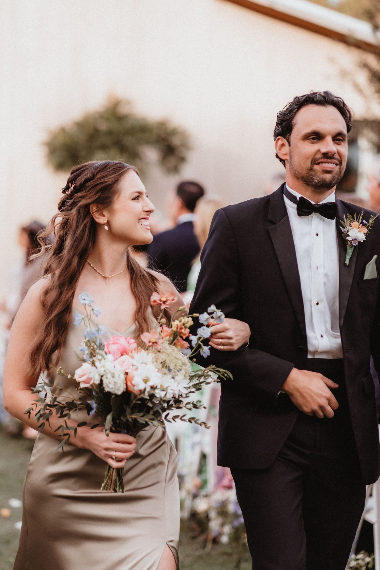 Smiling bridesmaid in gold dress holds white and pink bouquet while walking with groomsman in tuxedo.