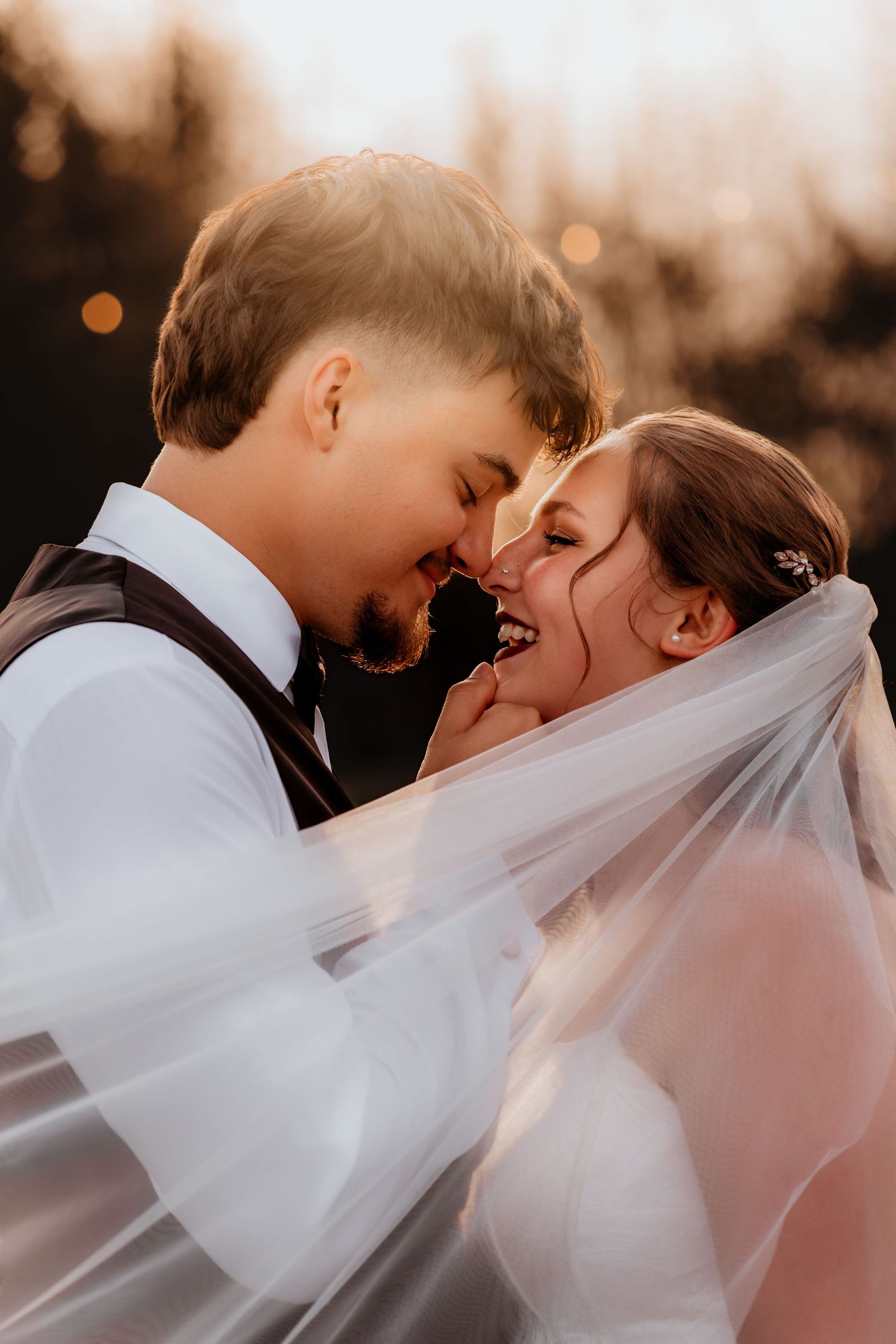 Joyful bride and groom nearly kissing beneath a flowing veil with warm bokeh sunset backdrop.