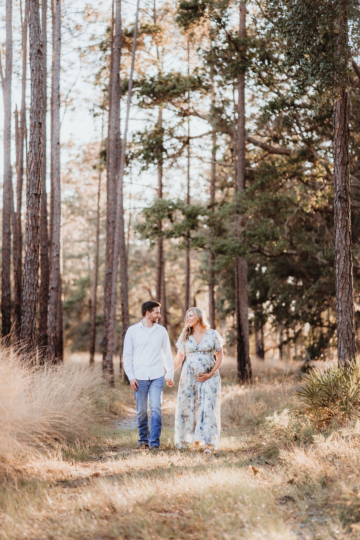 Expecting couple walking hand in hand through tall pine forest on sunny day.