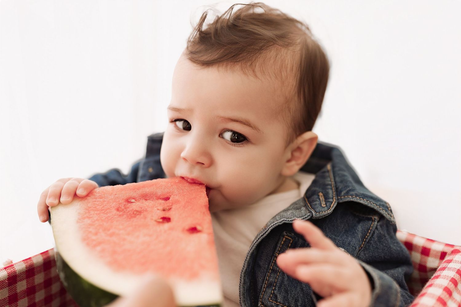 Adorable baby in denim jacket eating a fresh watermelon slice, enjoying a summer picnic.