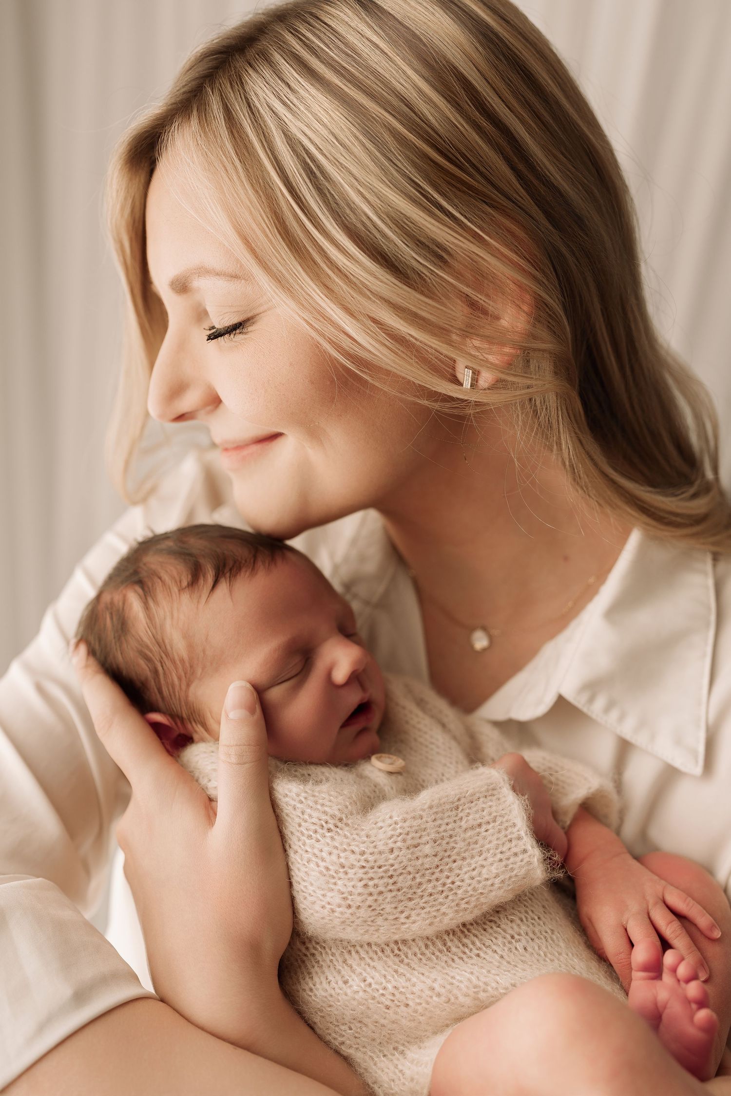 Blonde mother with closed eyes gently cradling her sleeping newborn baby in soft natural light.