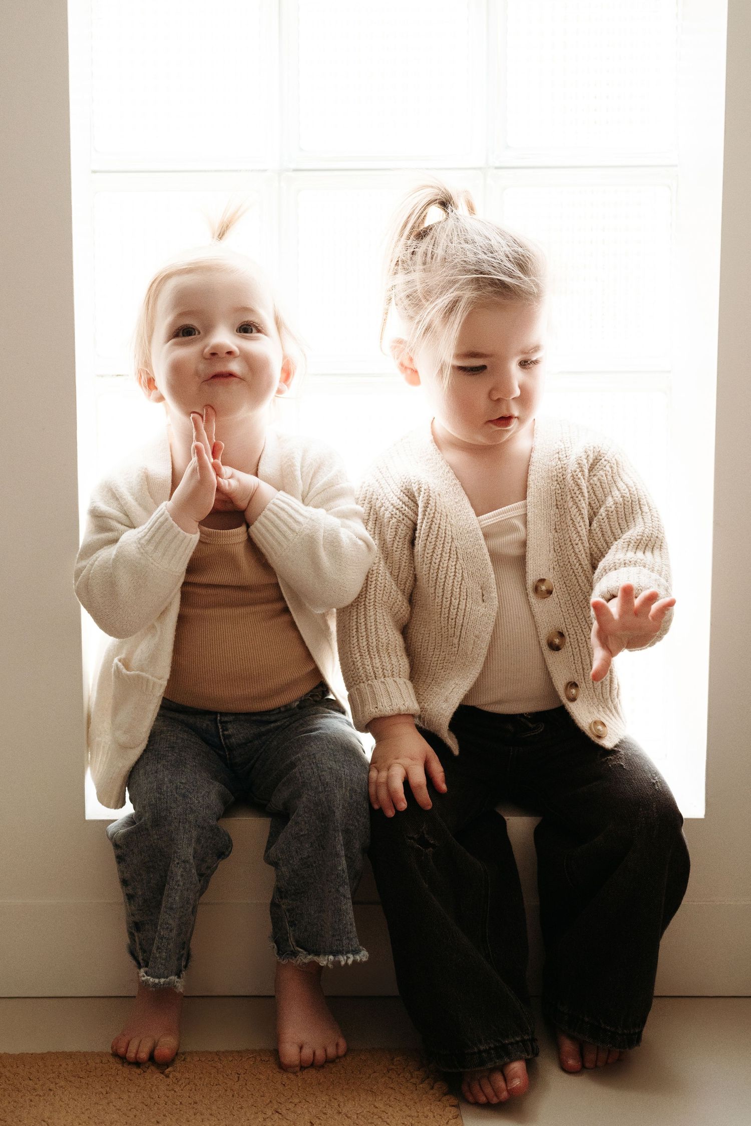 Two toddlers in cream knit cardigans sit barefoot on a window ledge, one clapping hands and smiling.