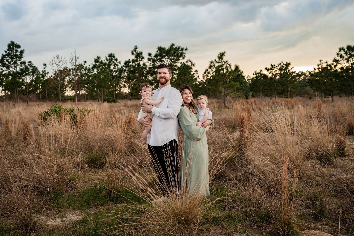 Family portrait of parents holding two babies in a rustic golden field at sunset.