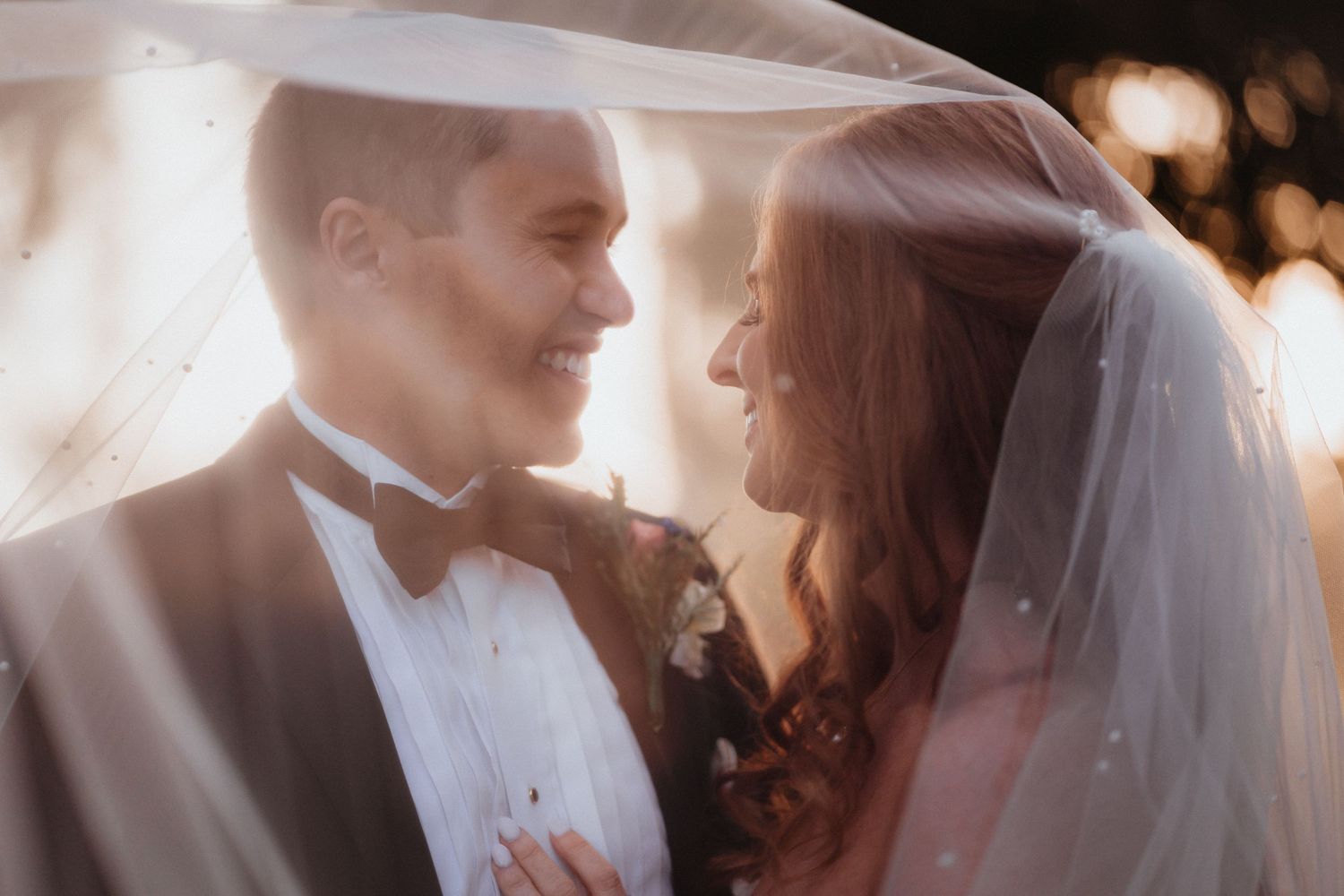 Bride and groom share a joyful moment under a pearl-dotted veil bathed in warm golden bokeh light.