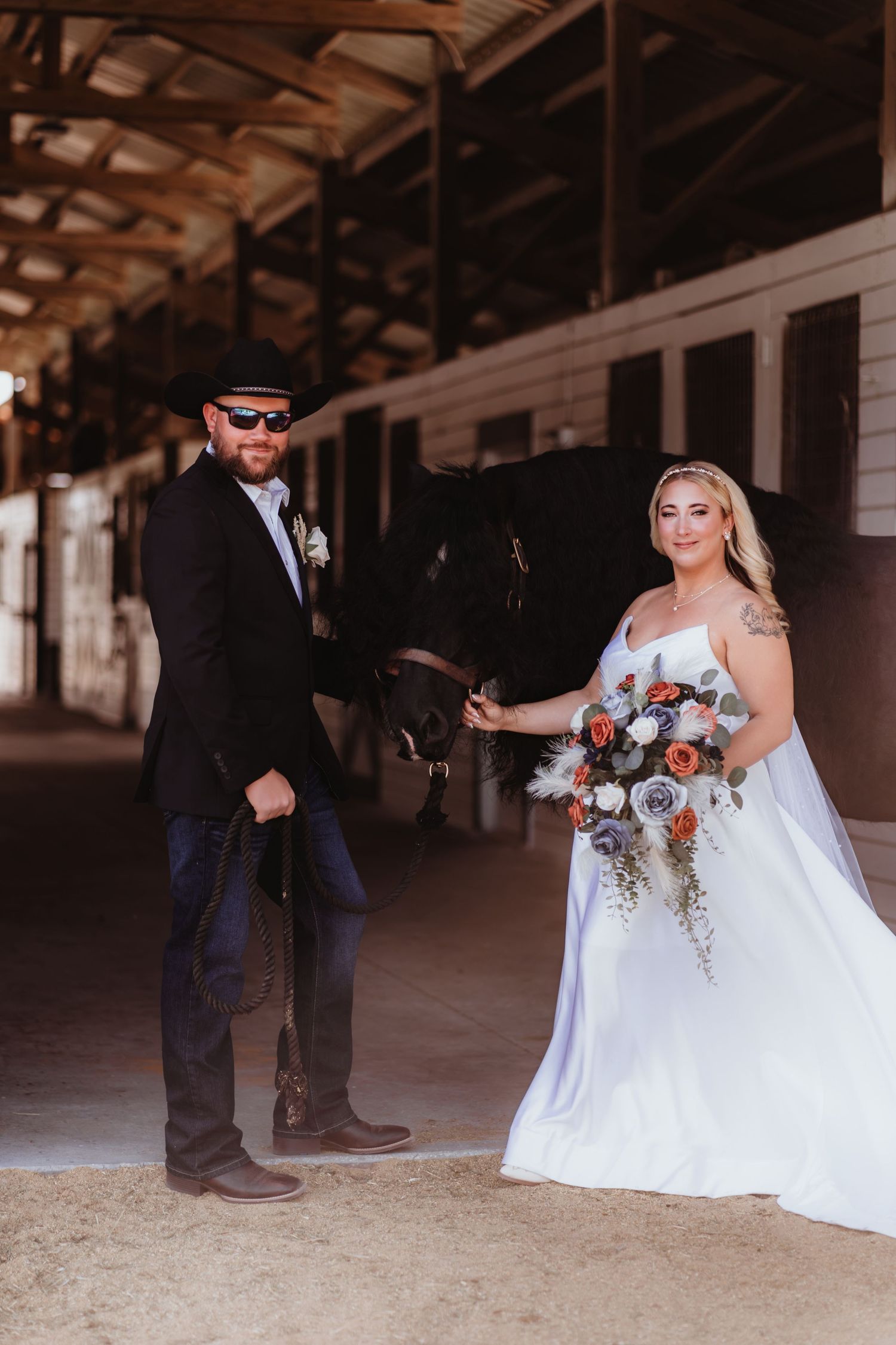 Bride and groom in cowboy hat posing with a black horse inside a rustic stable at their wedding.