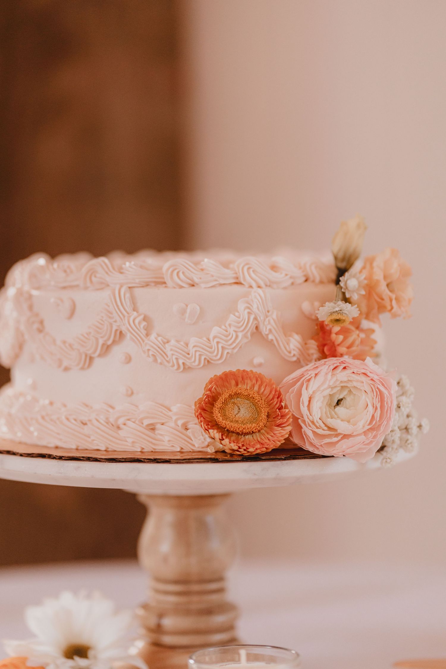 Elegant single-tier pink wedding cake with lace piping and fresh peach ranunculus flowers on wooden cake stand.