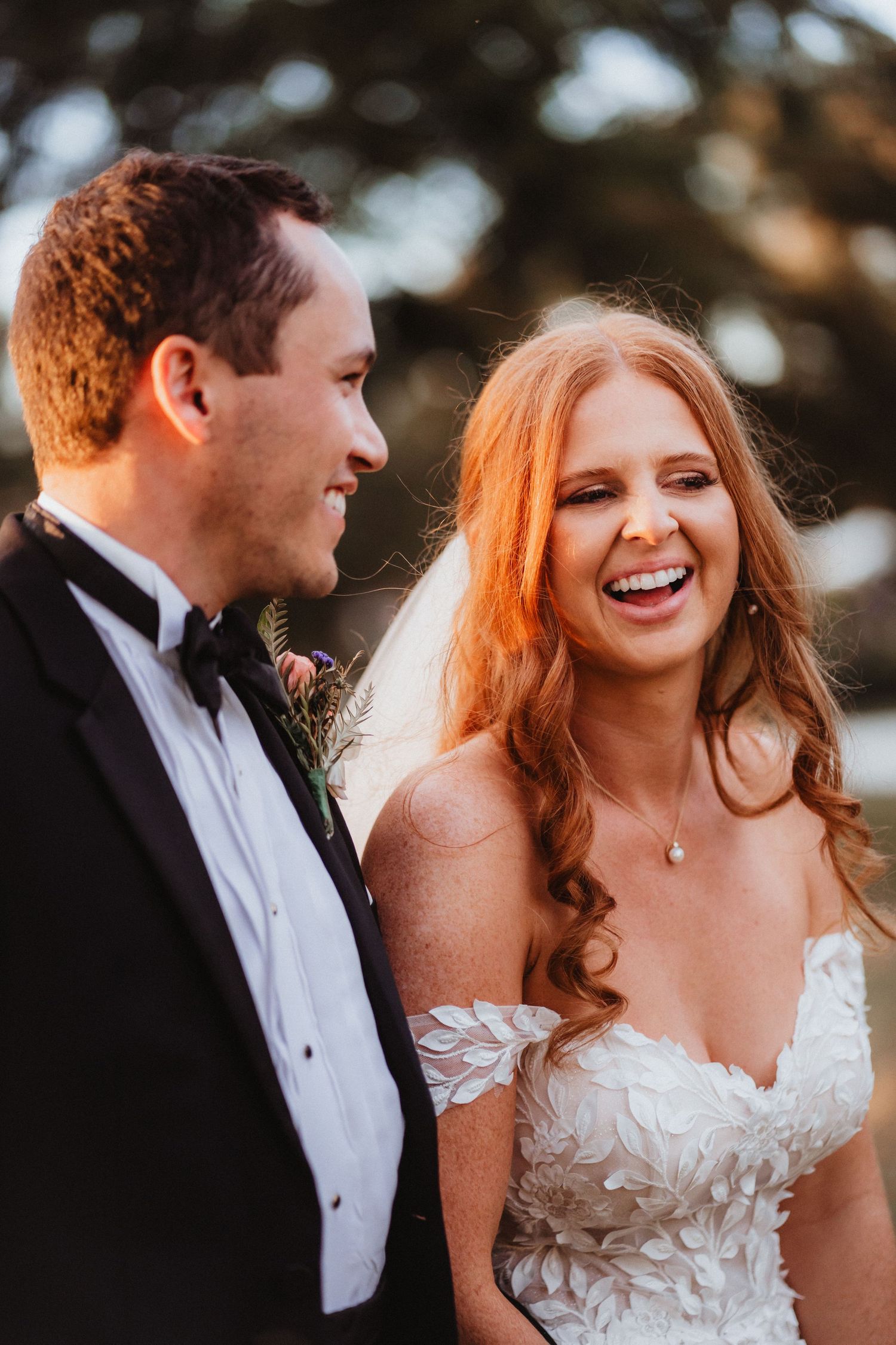 Close-up of joyful bride laughing beside groom in black tuxedo, bride wearing off-shoulder lace applique wedding gown.