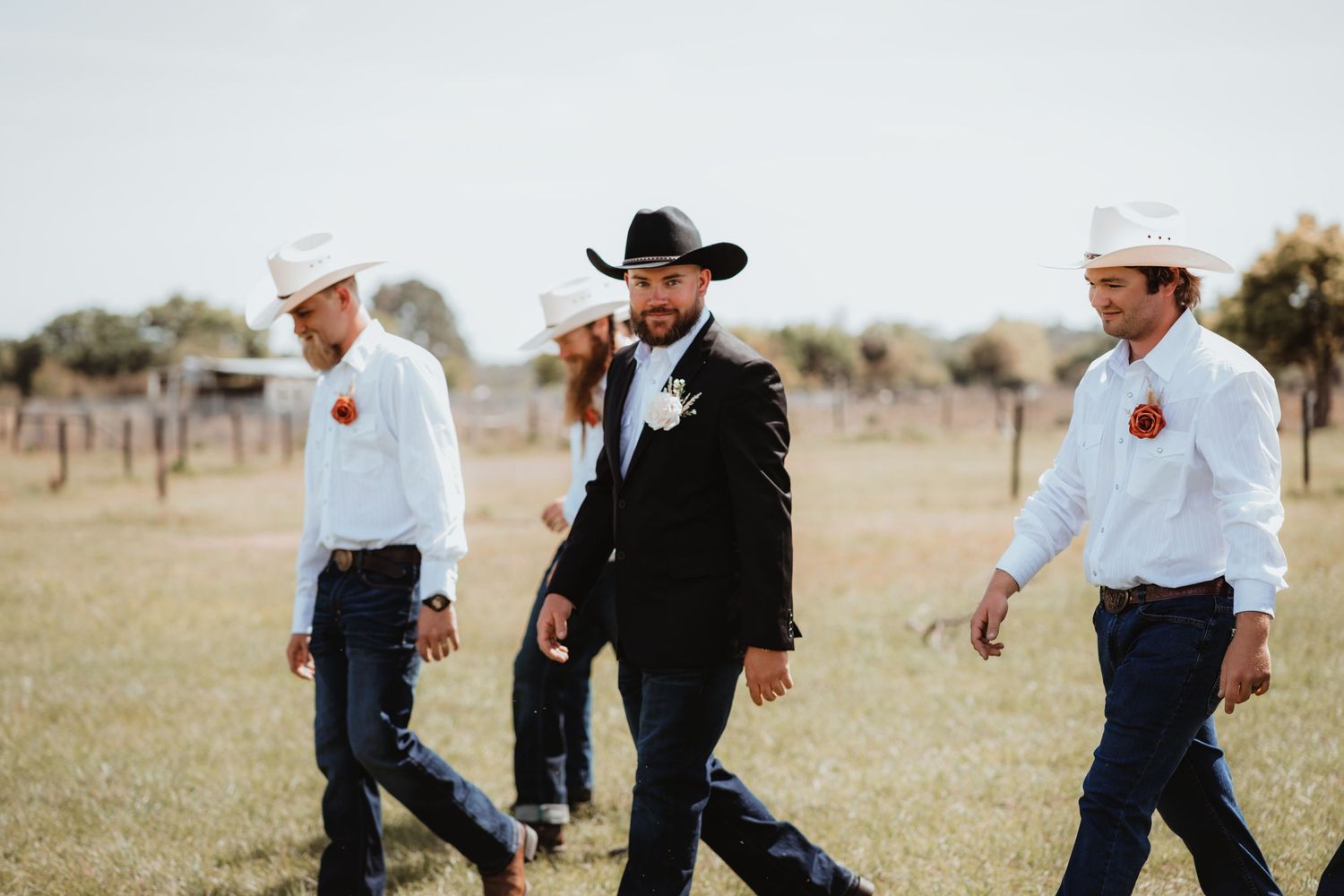 Groom in black hat and tuxedo strides with white-hatted groomsmen across an open ranch field.