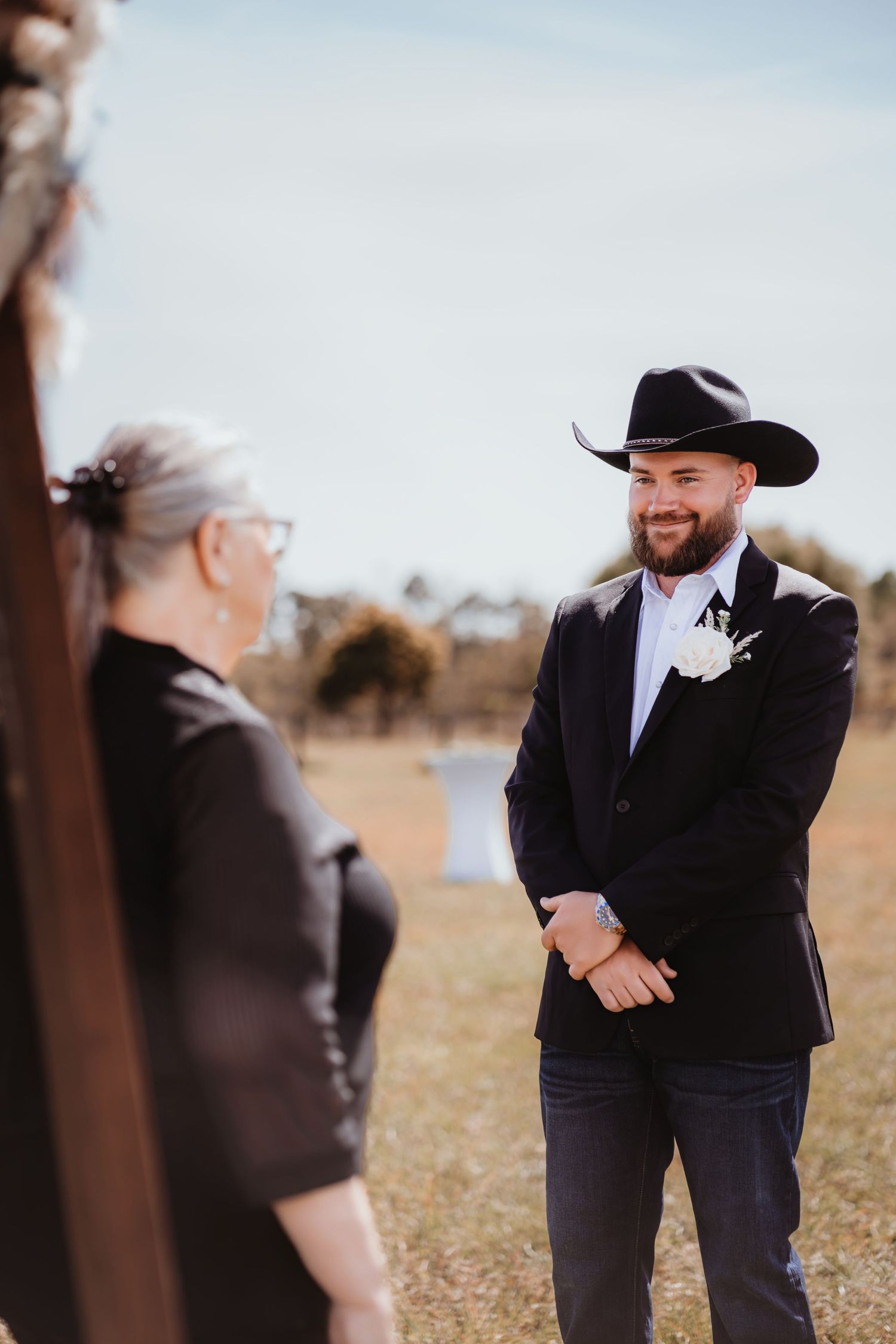 Smiling groom in black cowboy hat and boutonniere listens to officiant at outdoor rustic wedding ceremony.