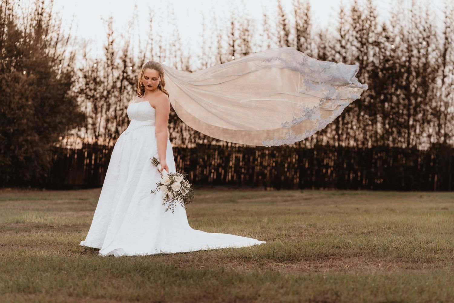 Bride in strapless white A-line gown with flowing veil holds bouquet in outdoor field.