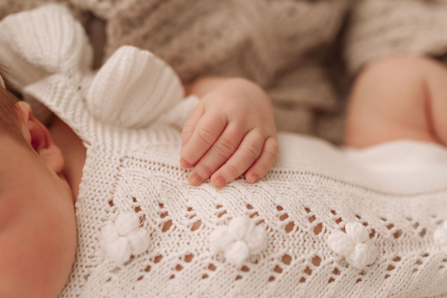 Close-up of a sleeping newborn baby's tiny hand resting on a white knitted blanket with delicate lace pattern.