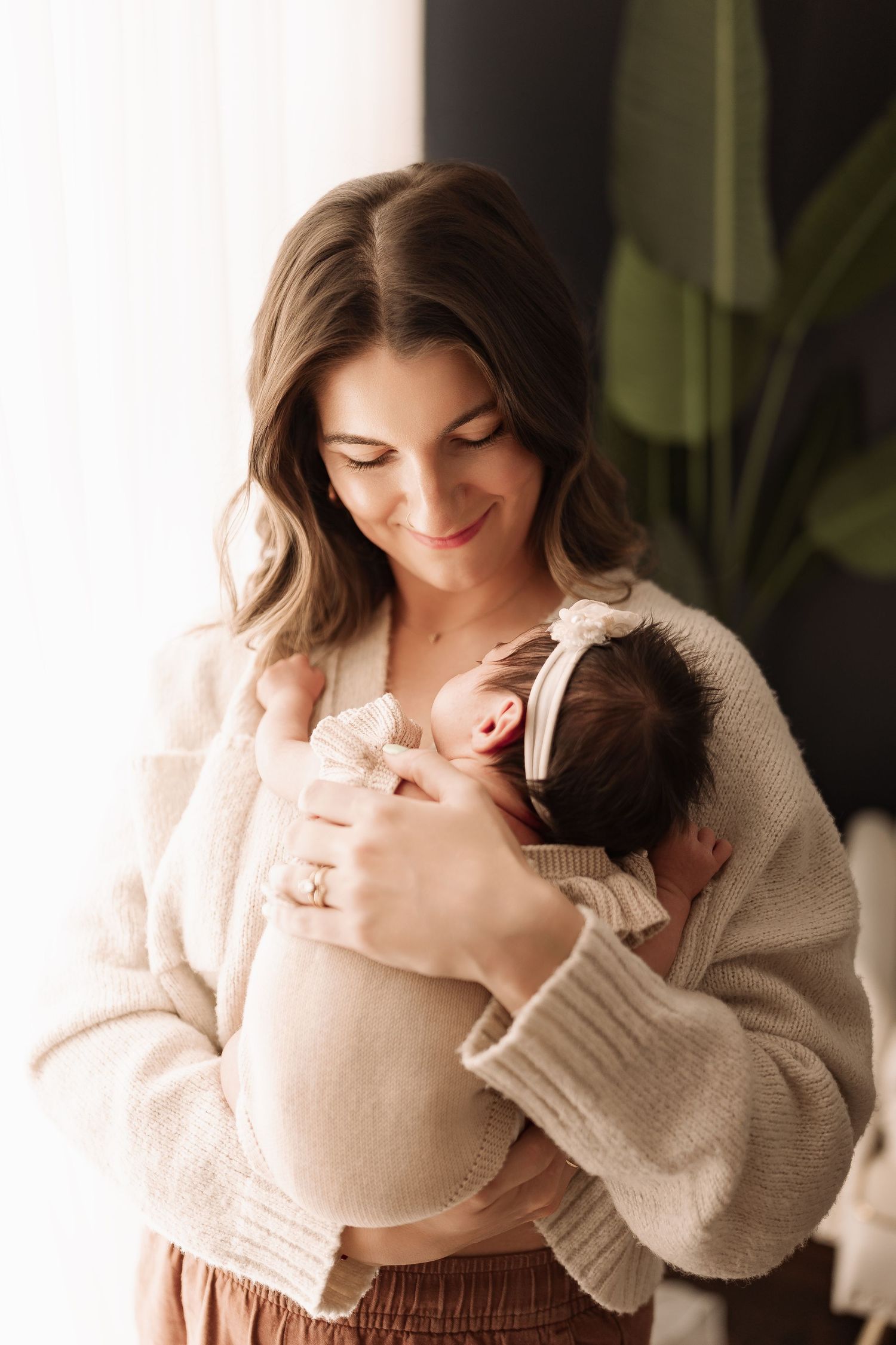 Smiling brunette mother in beige cardigan gently cradling sleeping newborn with pink bow headband.